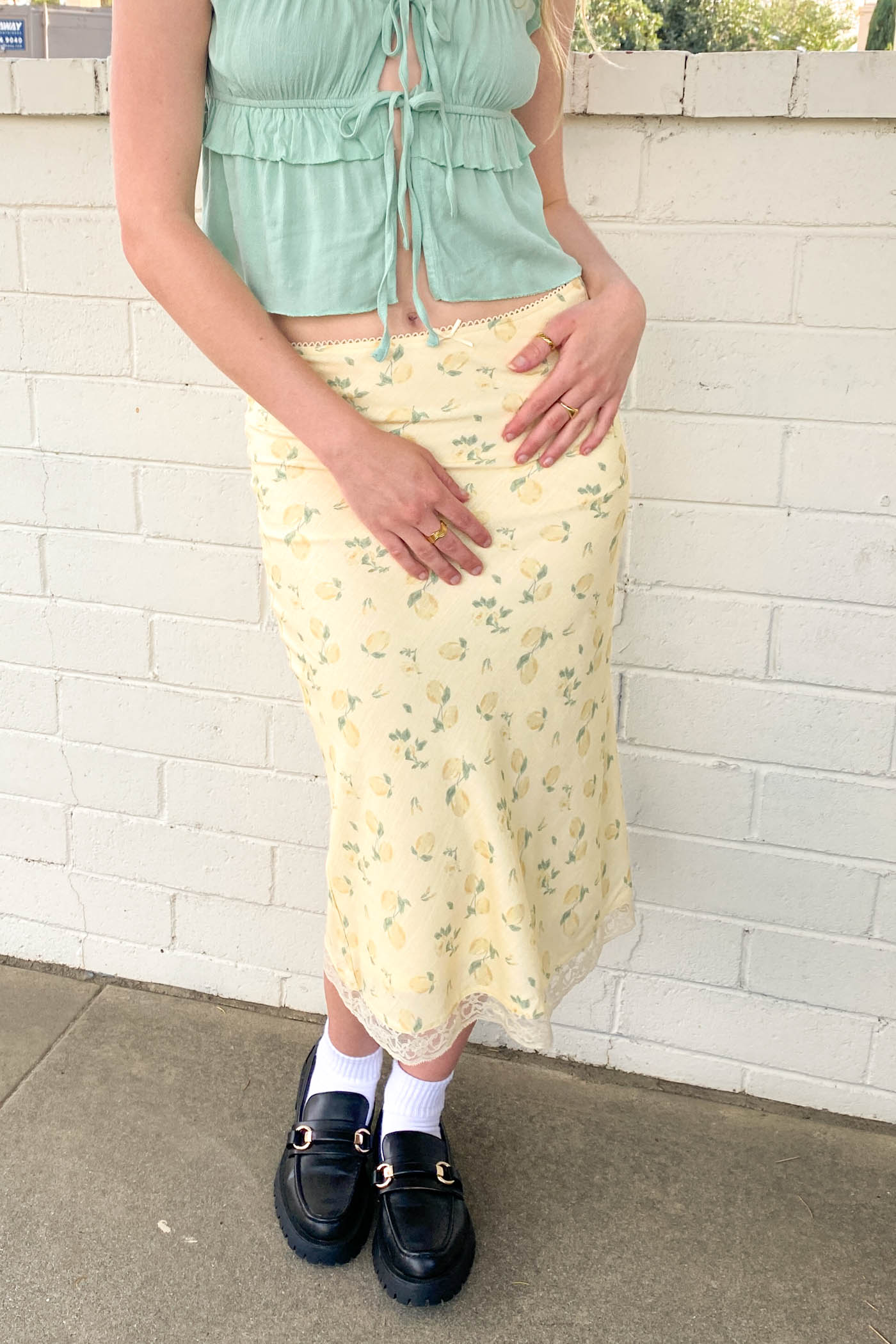 Person wearing a light green top and yellow floral skirt with black shoes against a white brick wall.