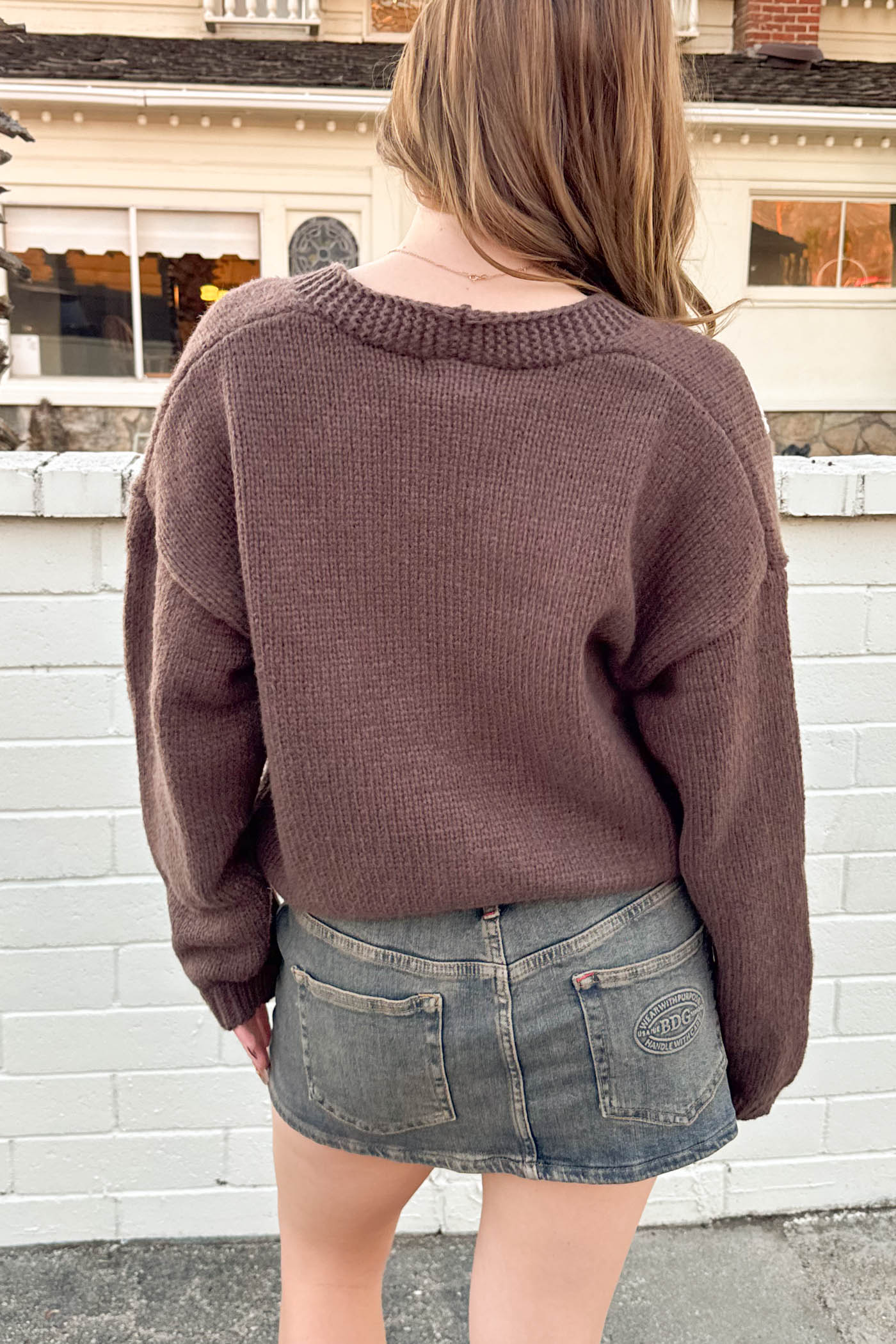 Person wearing a brown sweater and denim skirt standing in front of a white brick wall.