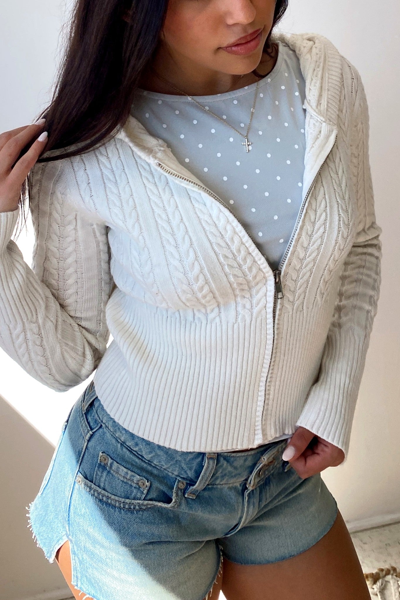 Woman wearing a light blue polka dot top, cream cardigan, and denim shorts indoors.
