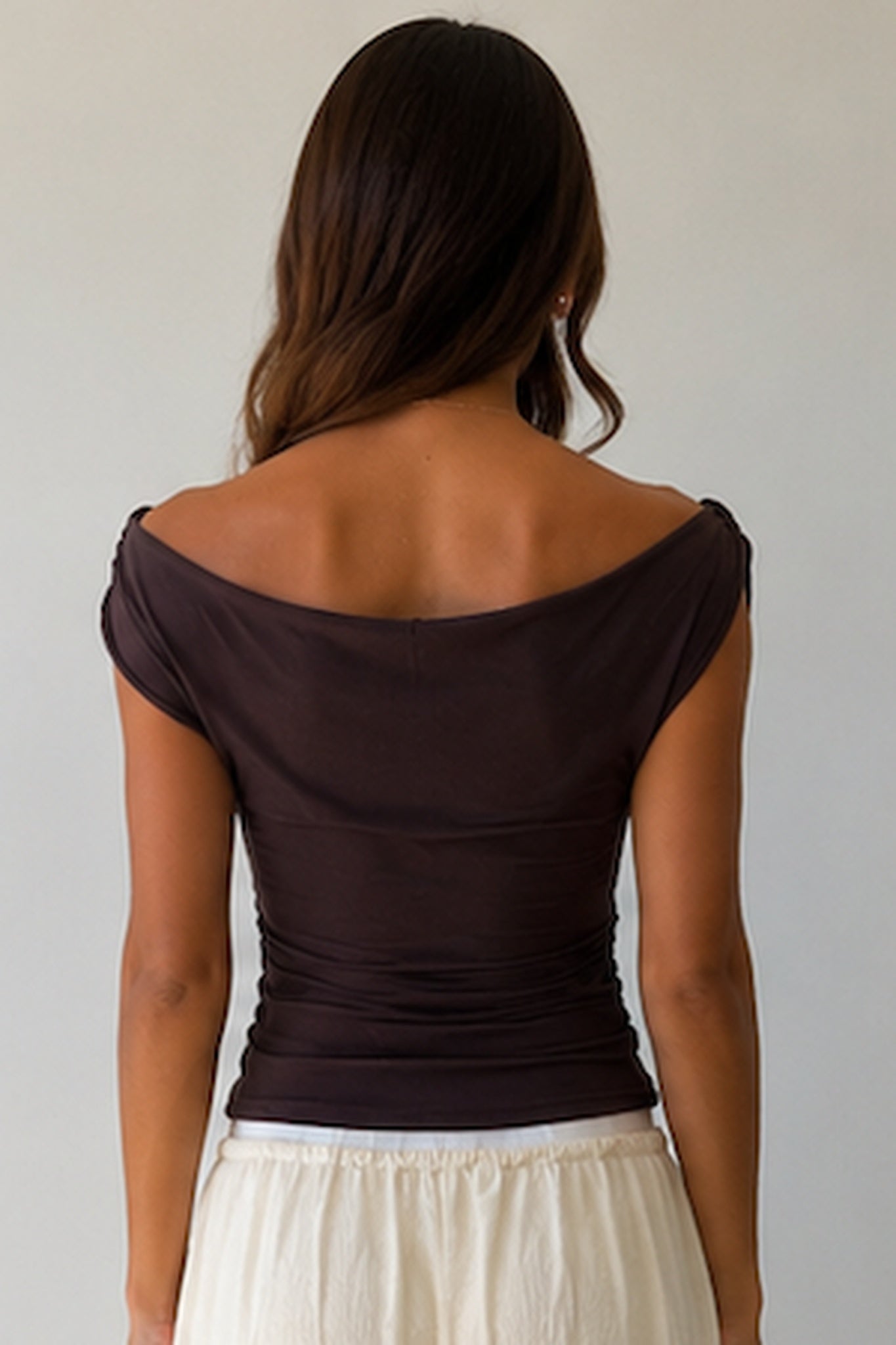 A woman viewed from behind wearing a dark brown off-the-shoulder short-sleeve top and a cream skirt, set against a plain light gray background.