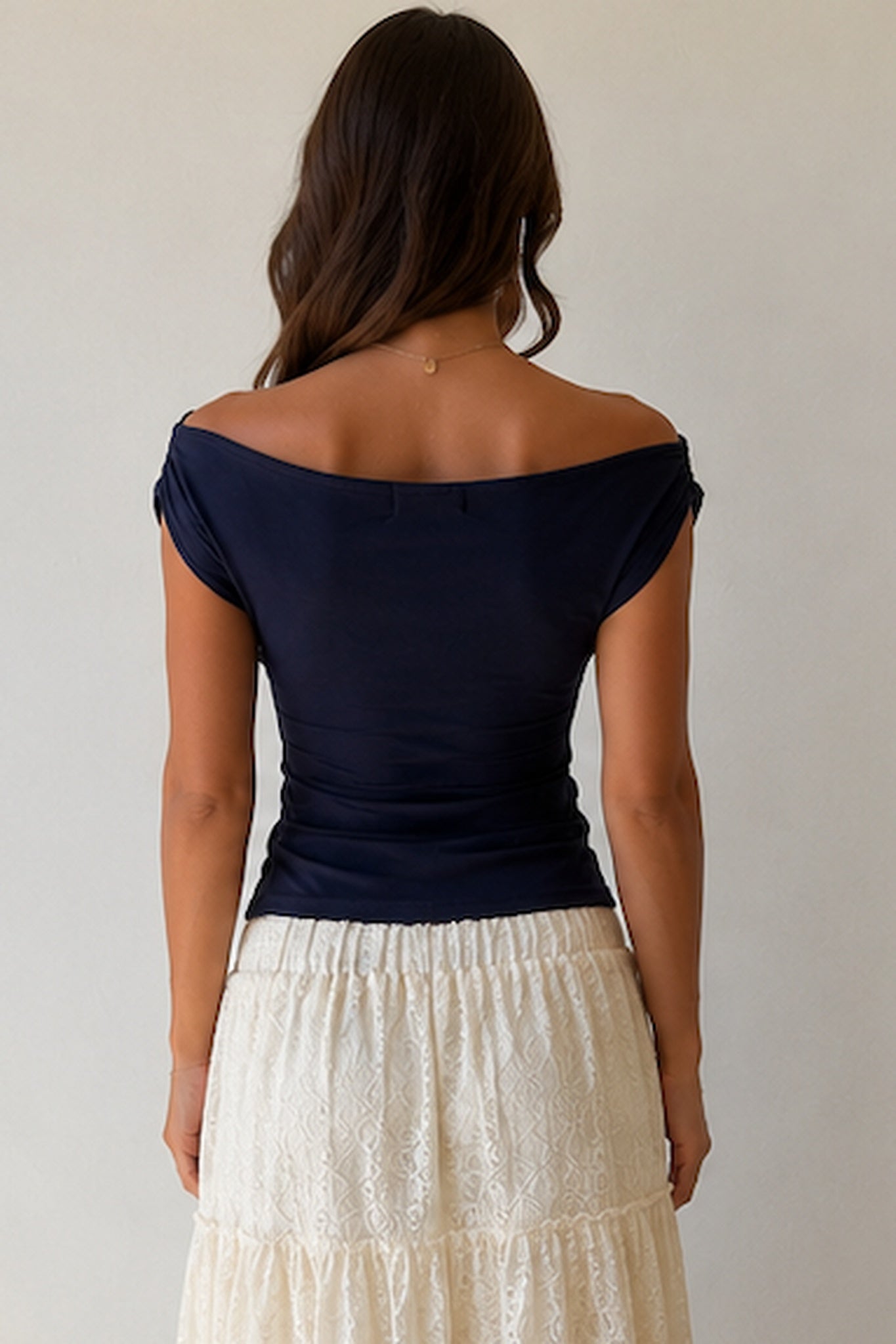 A woman wearing a navy blue off-the-shoulder top with ruched detailing at the waist, paired with a cream-colored textured skirt. She is wearing a delicate gold necklace.