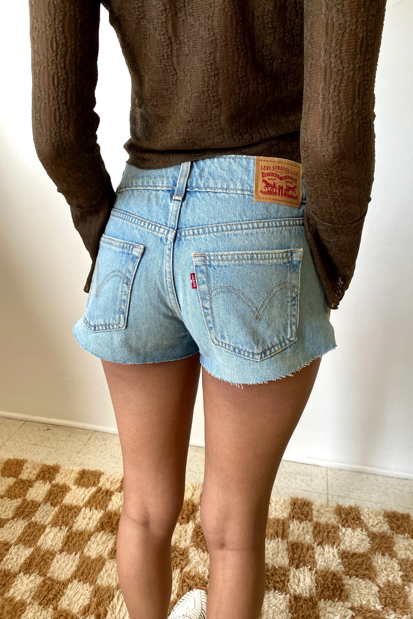 Person wearing a brown sweater and light blue denim shorts with a visible brand logo on a checkered floor.