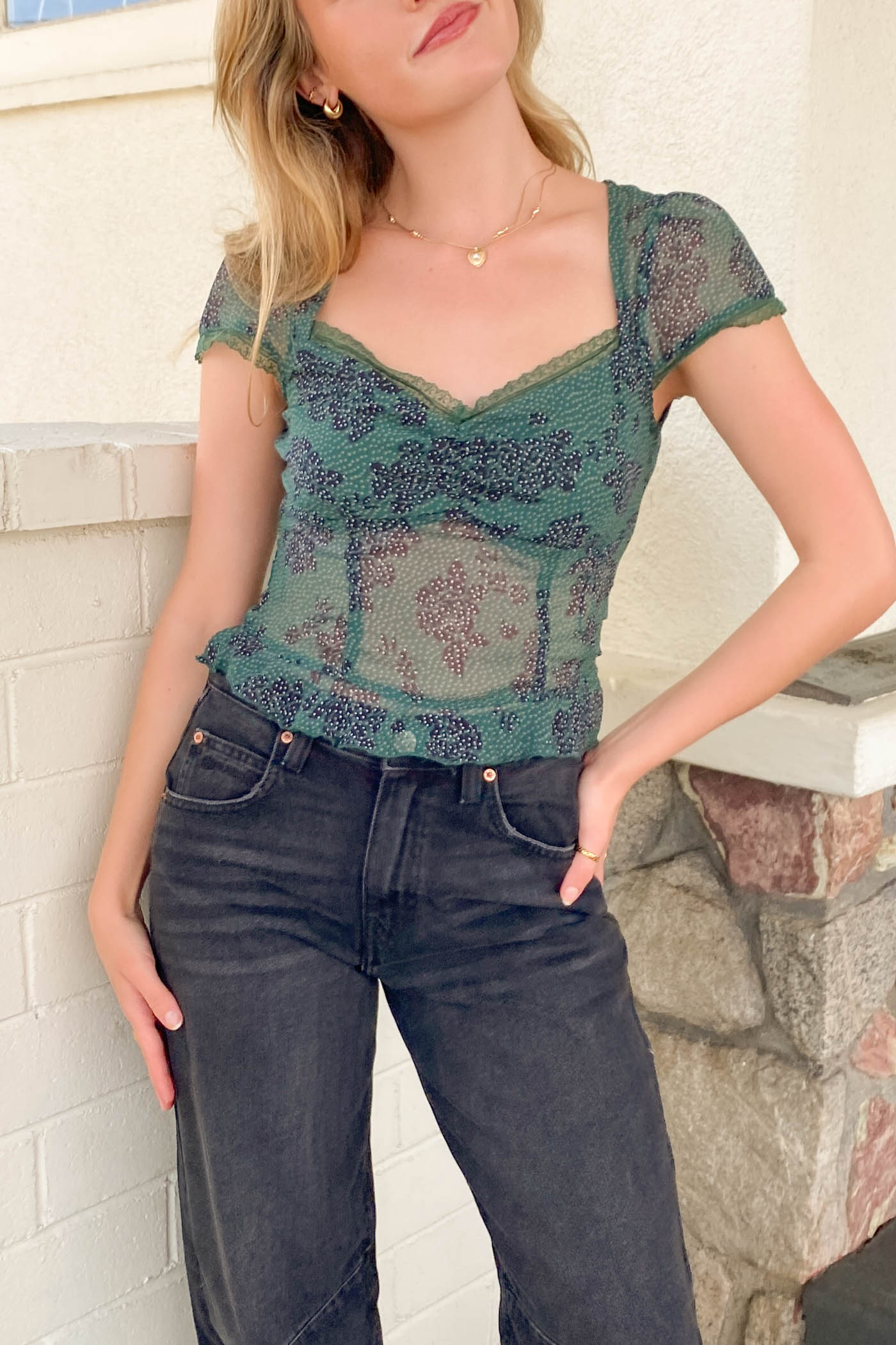 Woman wearing a green lace top and dark jeans against a stone wall.