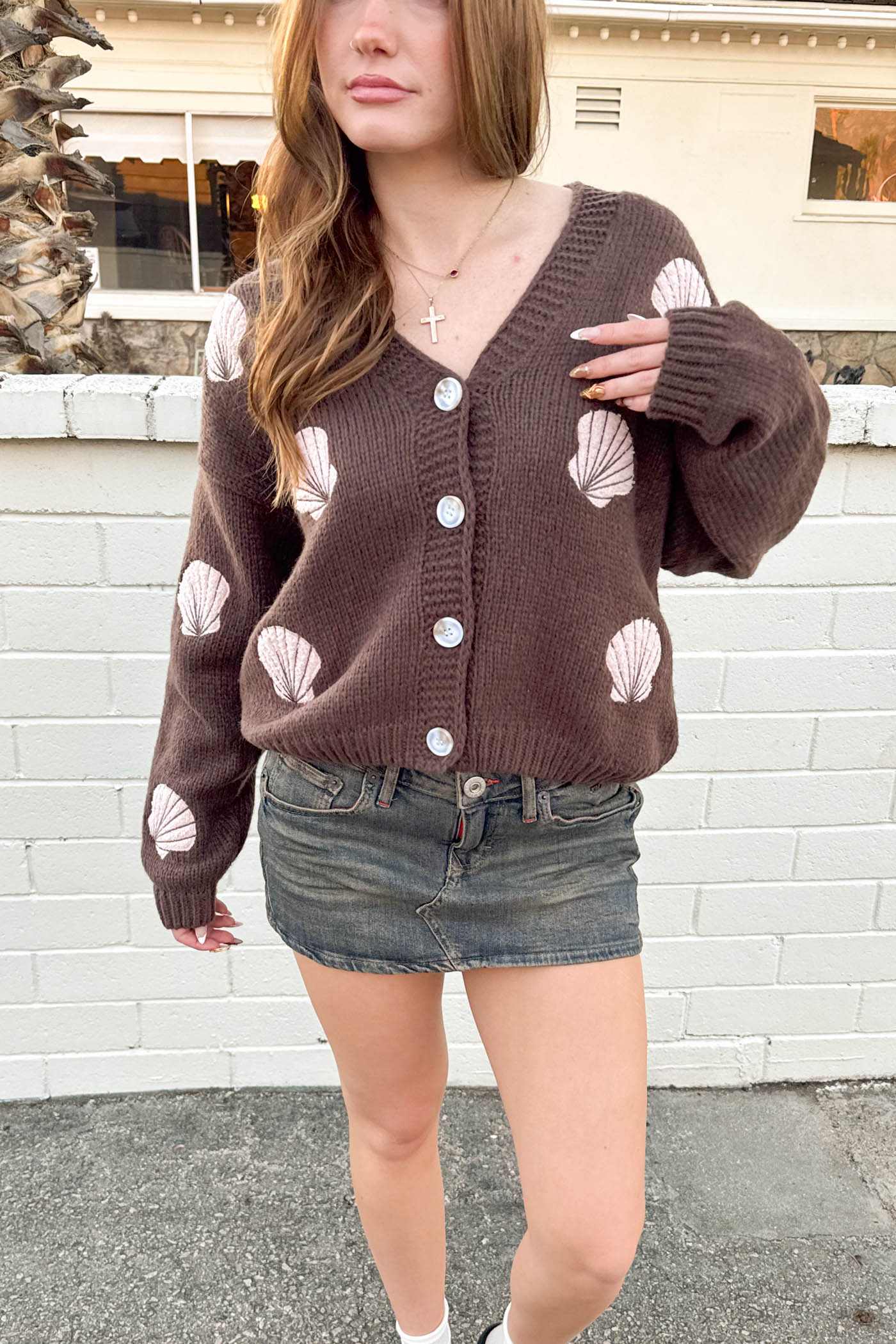 Person wearing a brown cardigan with shell patterns in front of a white brick wall.