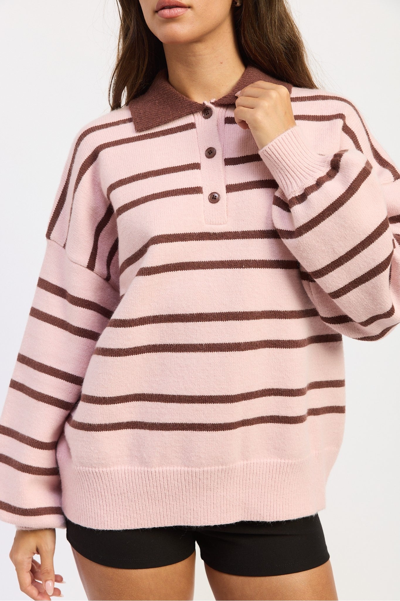 A woman wearing a pink and brown horizontally striped knit polo sweater with a three-button placket, paired with black shorts, against a plain white background.