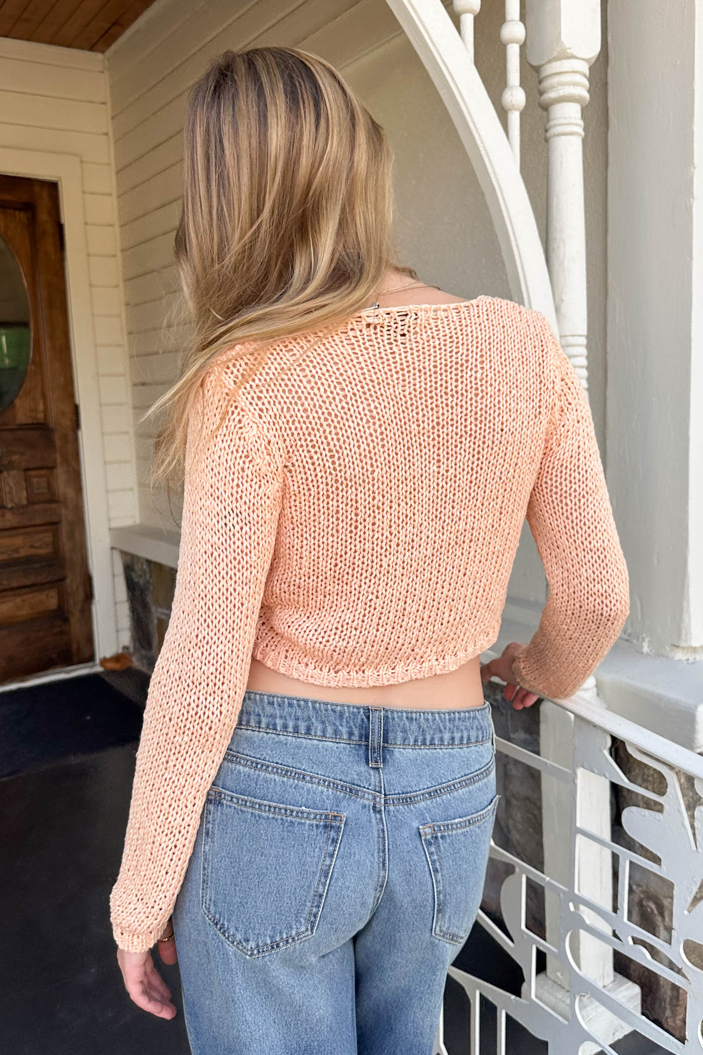 Person wearing a peach knitted top and blue jeans standing on a porch.