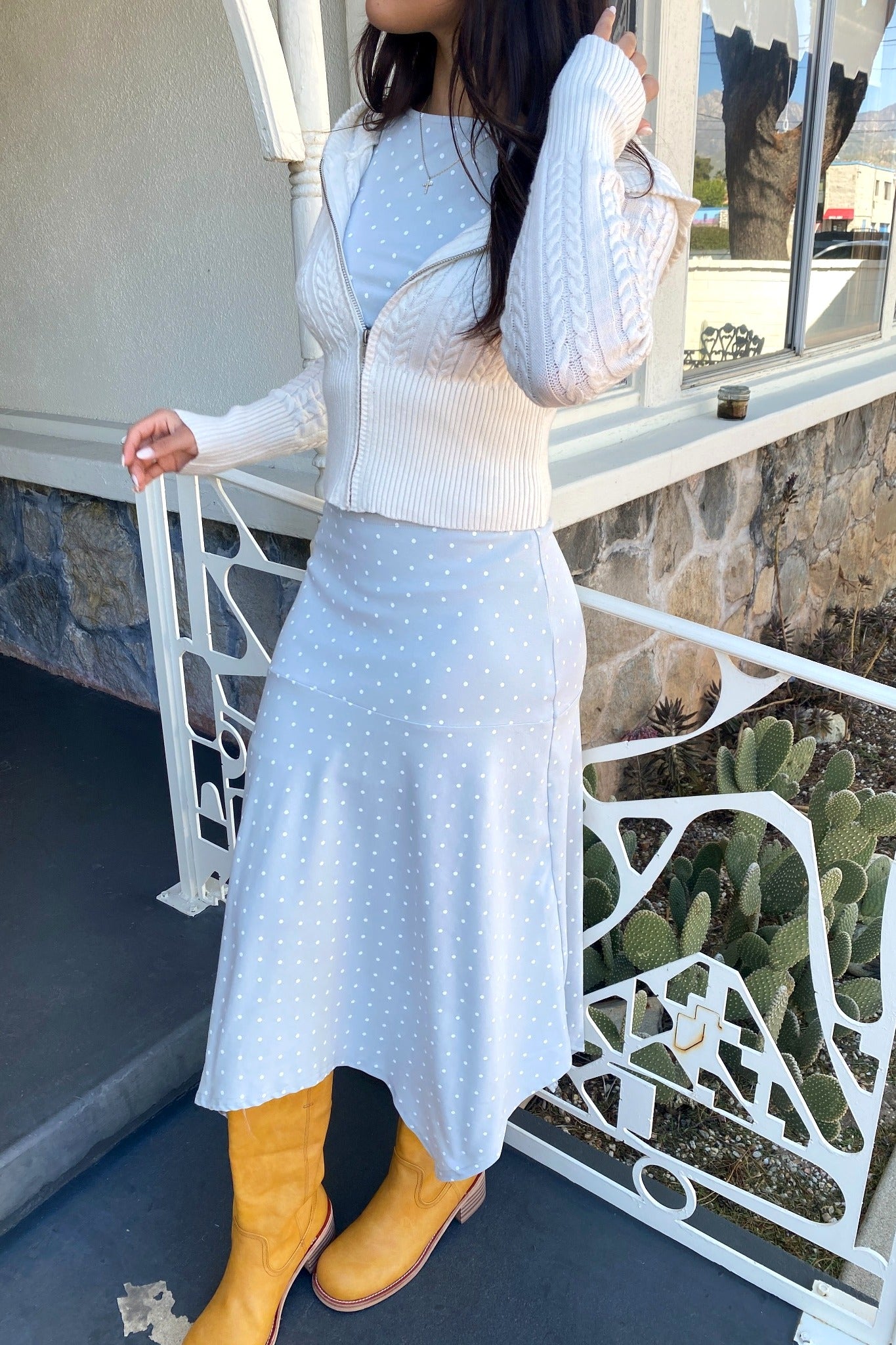 Person wearing a light blue dress with white polka dots and yellow boots, standing on a balcony.