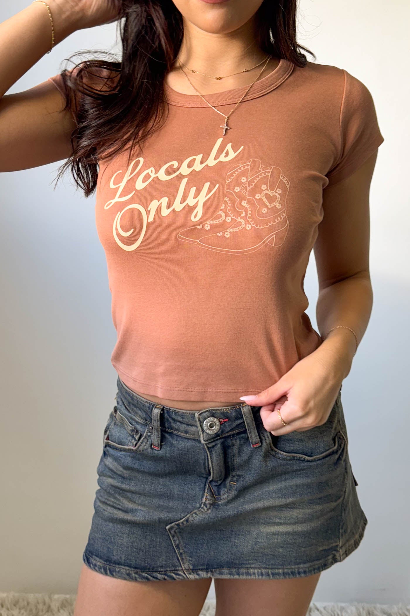 Person wearing a brown t-shirt with 'Locals Only' text and a denim skirt against a plain background
