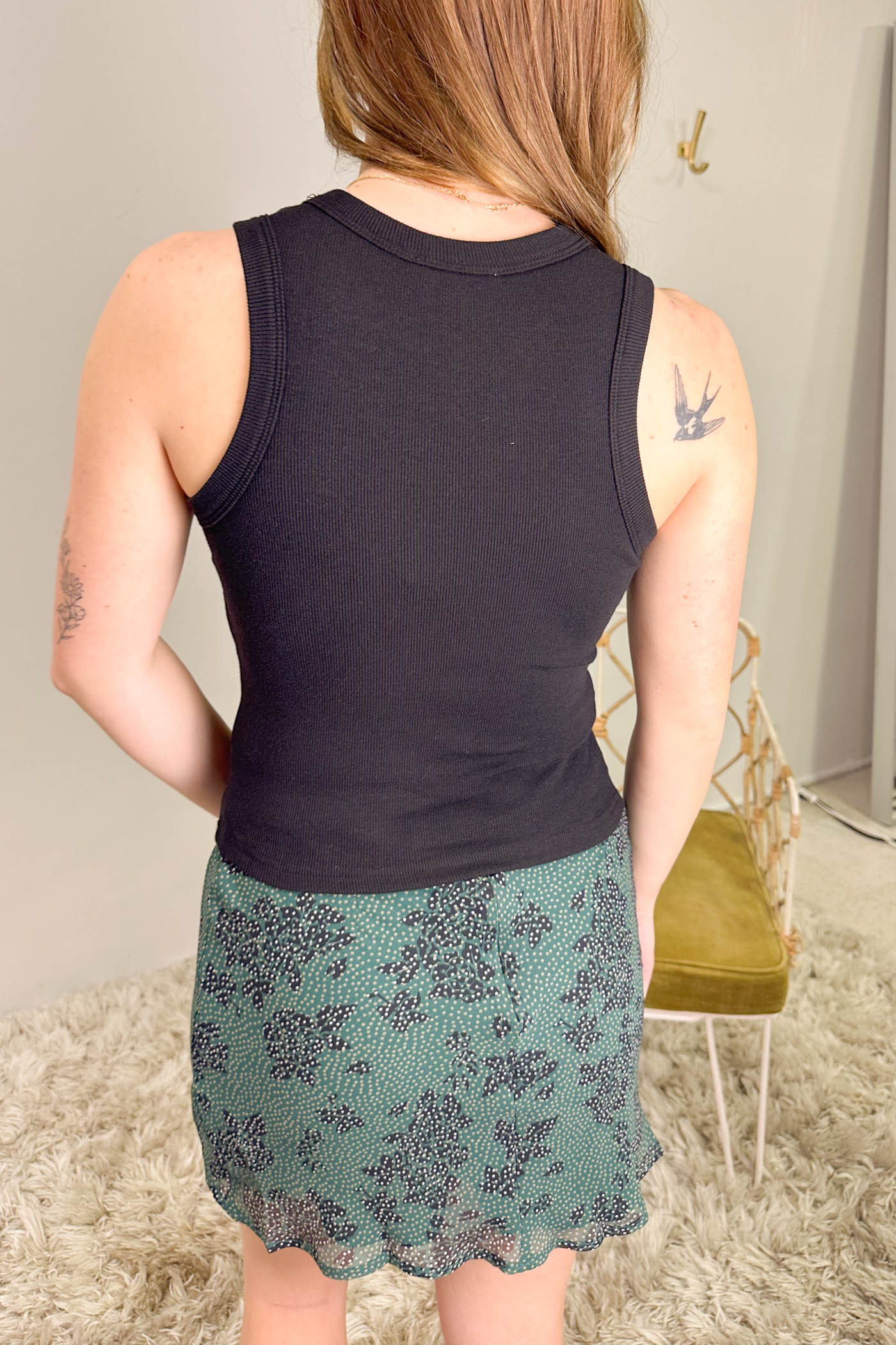 Person wearing a black tank top and green floral skirt in a room with a chair and coat rack.