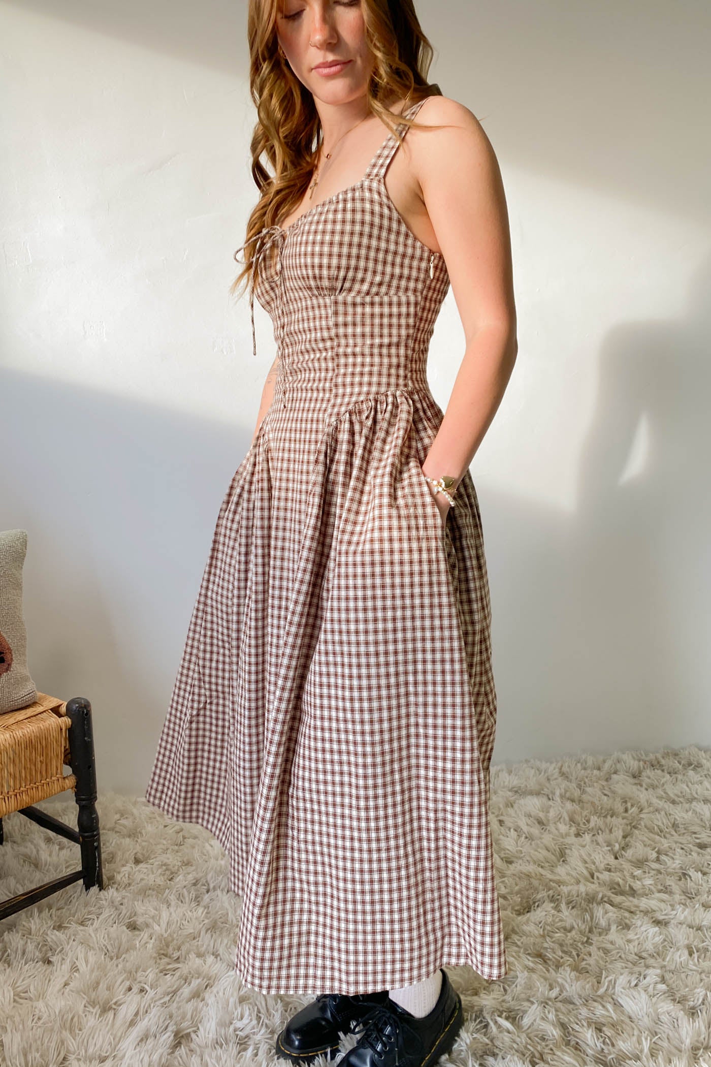 Woman wearing a checkered dress standing in a room with a white wall and light-colored carpet.