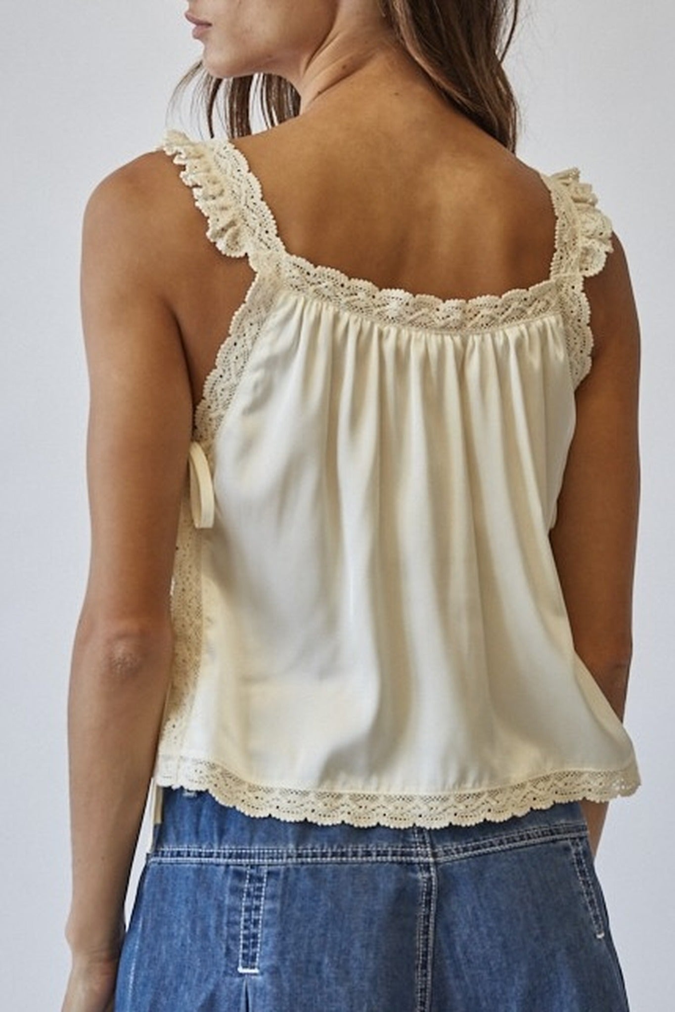 Woman wearing a cream-colored tank top with lace details and blue jeans on a plain background