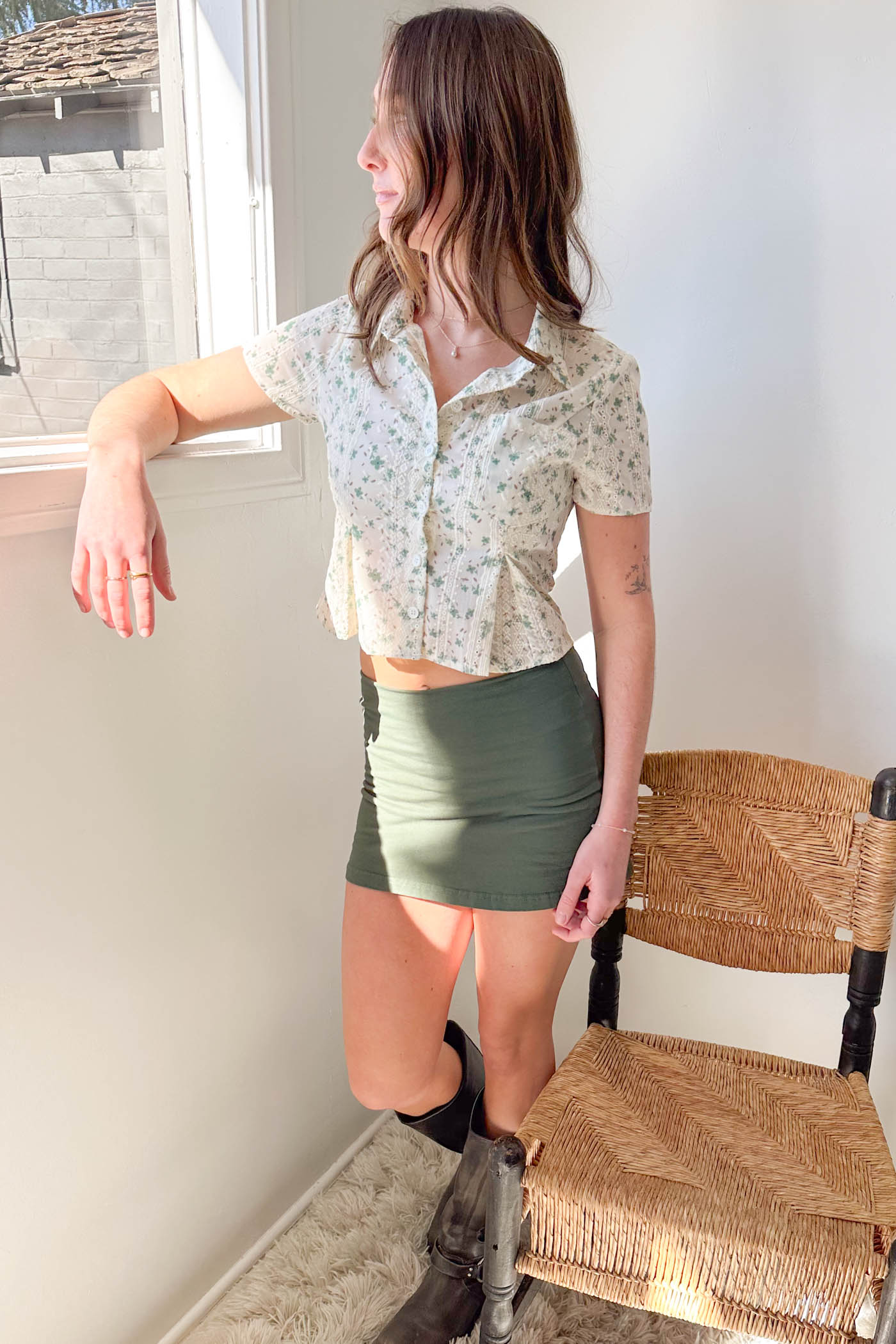 Woman standing indoors wearing a floral blouse and green skirt, leaning against a chair.