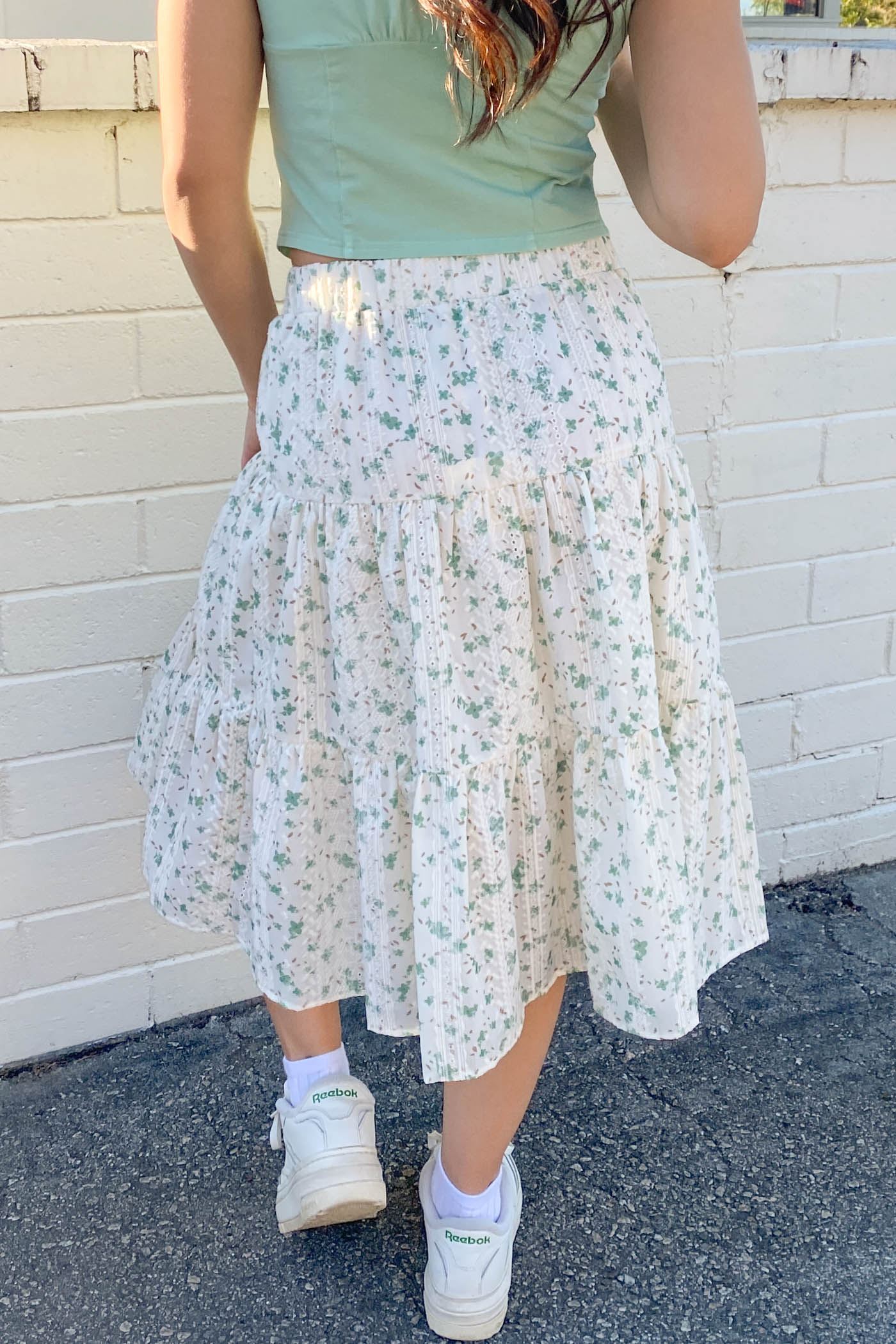 Person wearing a light green top and floral skirt against a white brick wall.