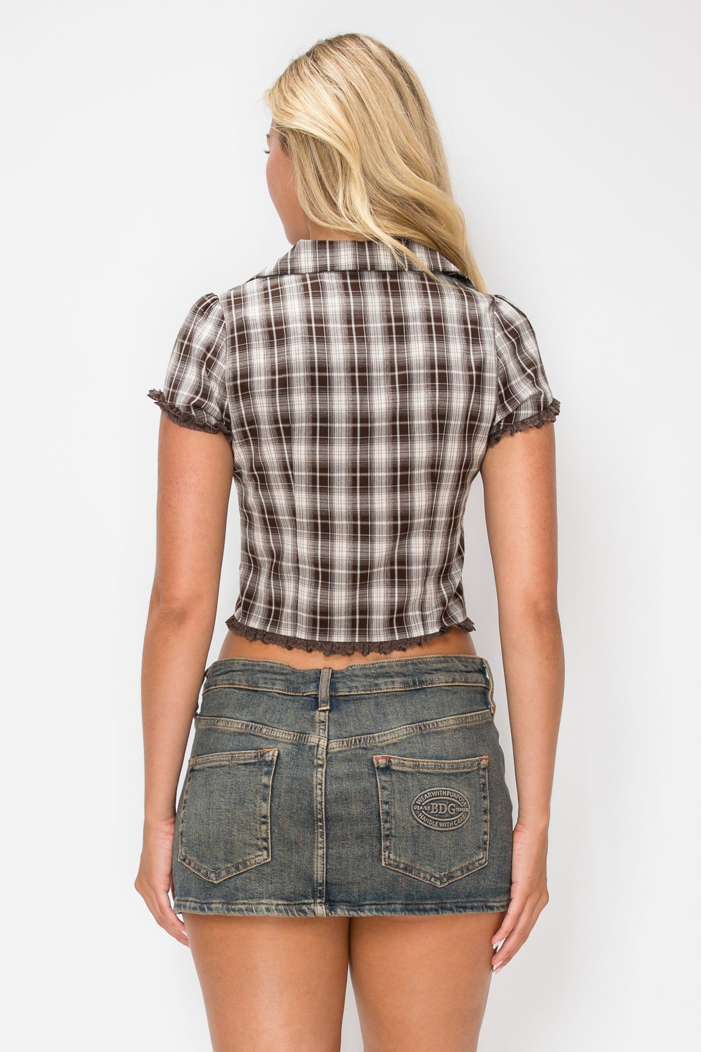 A woman shown from the back wearing a brown and beige plaid short-sleeve cropped shirt with ruffled trim, paired with blue denim shorts, against a light gray studio background.