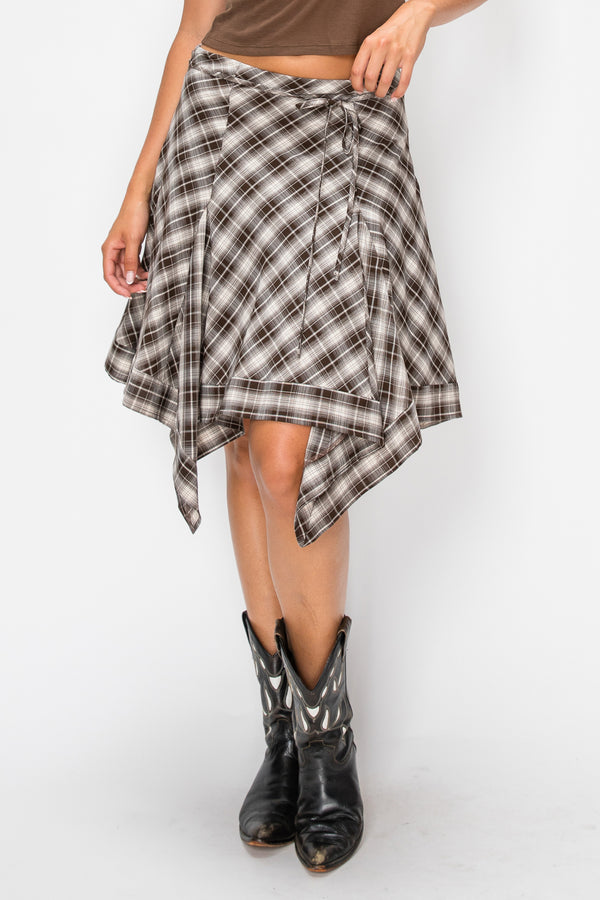 A woman wearing a brown, black, and white plaid wrap skirt with an asymmetrical hem, styled with black cowboy boots against a plain white studio background.
