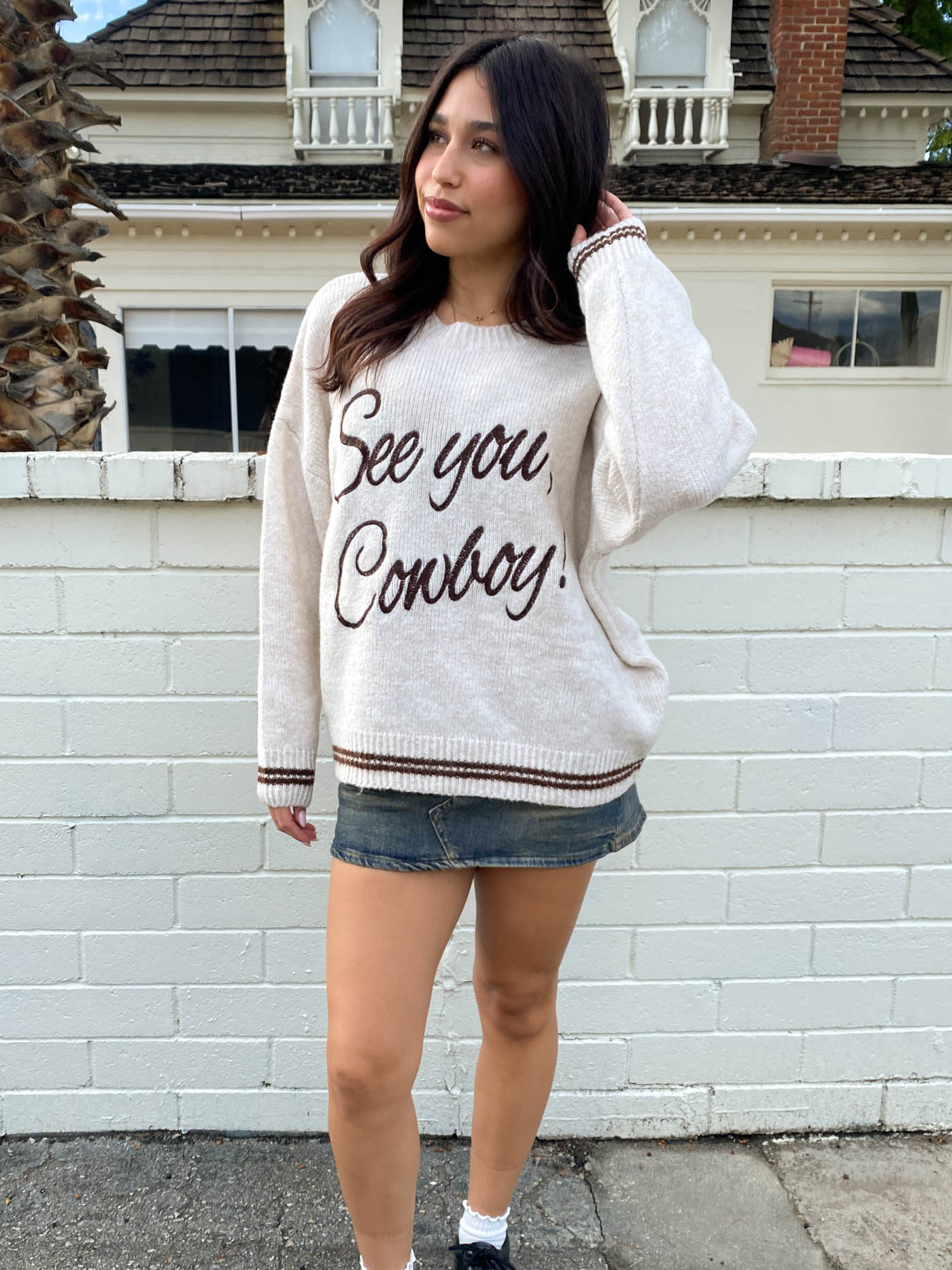 Woman wearing a sweater with 'See you, Cowboy!' text in front of a white brick wall.