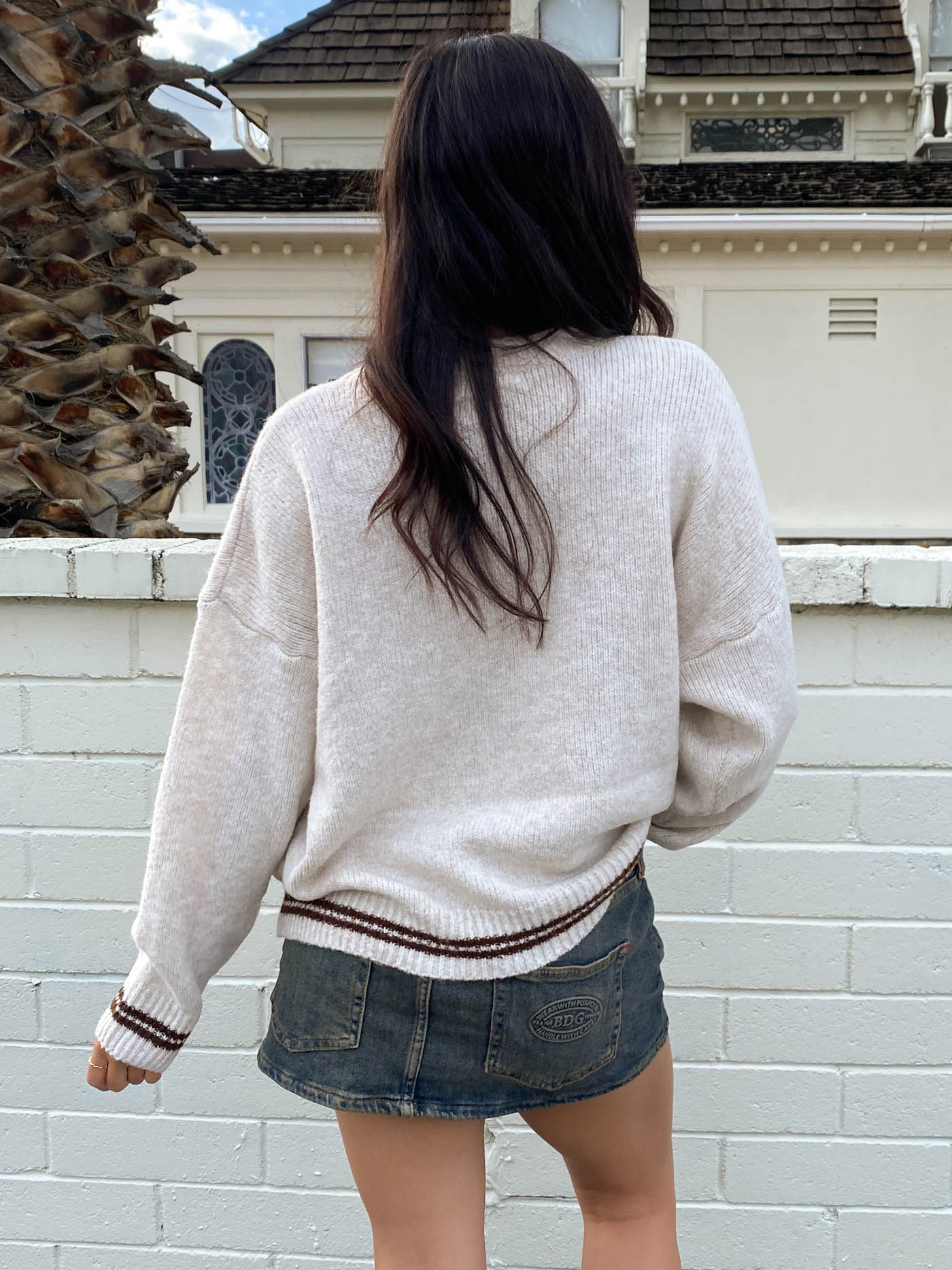 Person wearing a light-colored sweater and denim skirt standing in front of a white brick wall.