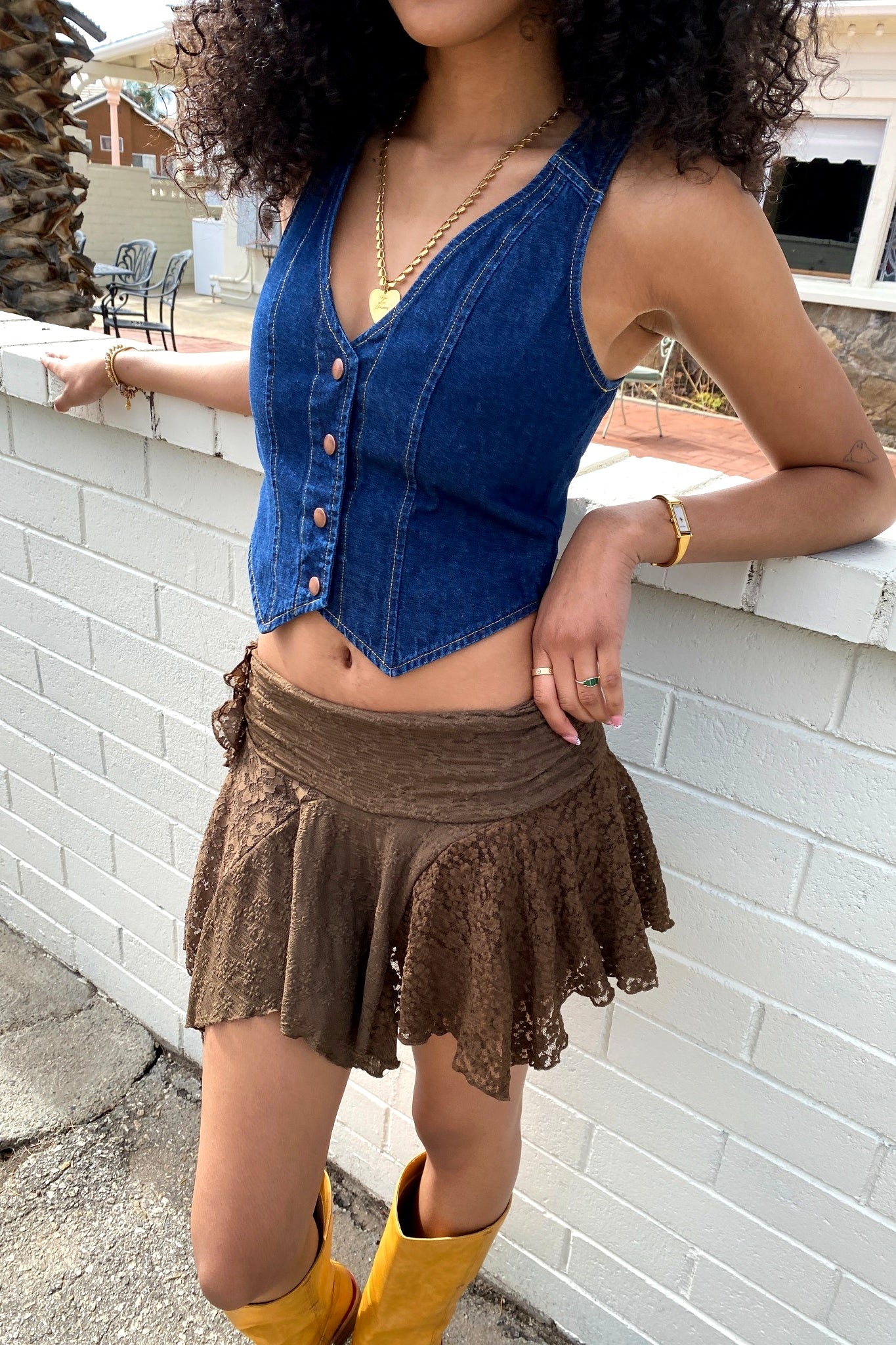 Woman wearing a blue denim vest over a brown lace skirt with yellow boots, leaning against a white brick wall.