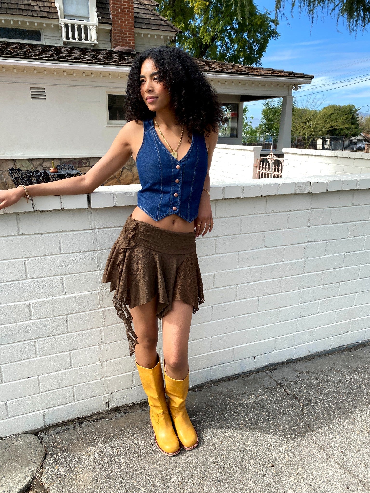 Woman in blue top and brown skirt with yellow boots standing against a white wall.