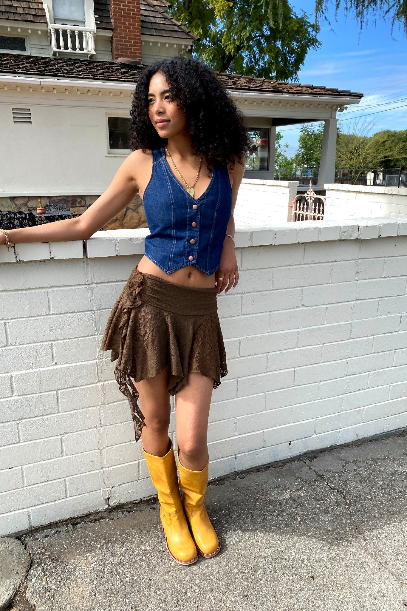 Woman in a blue top and brown skirt with yellow boots standing against a white brick wall.