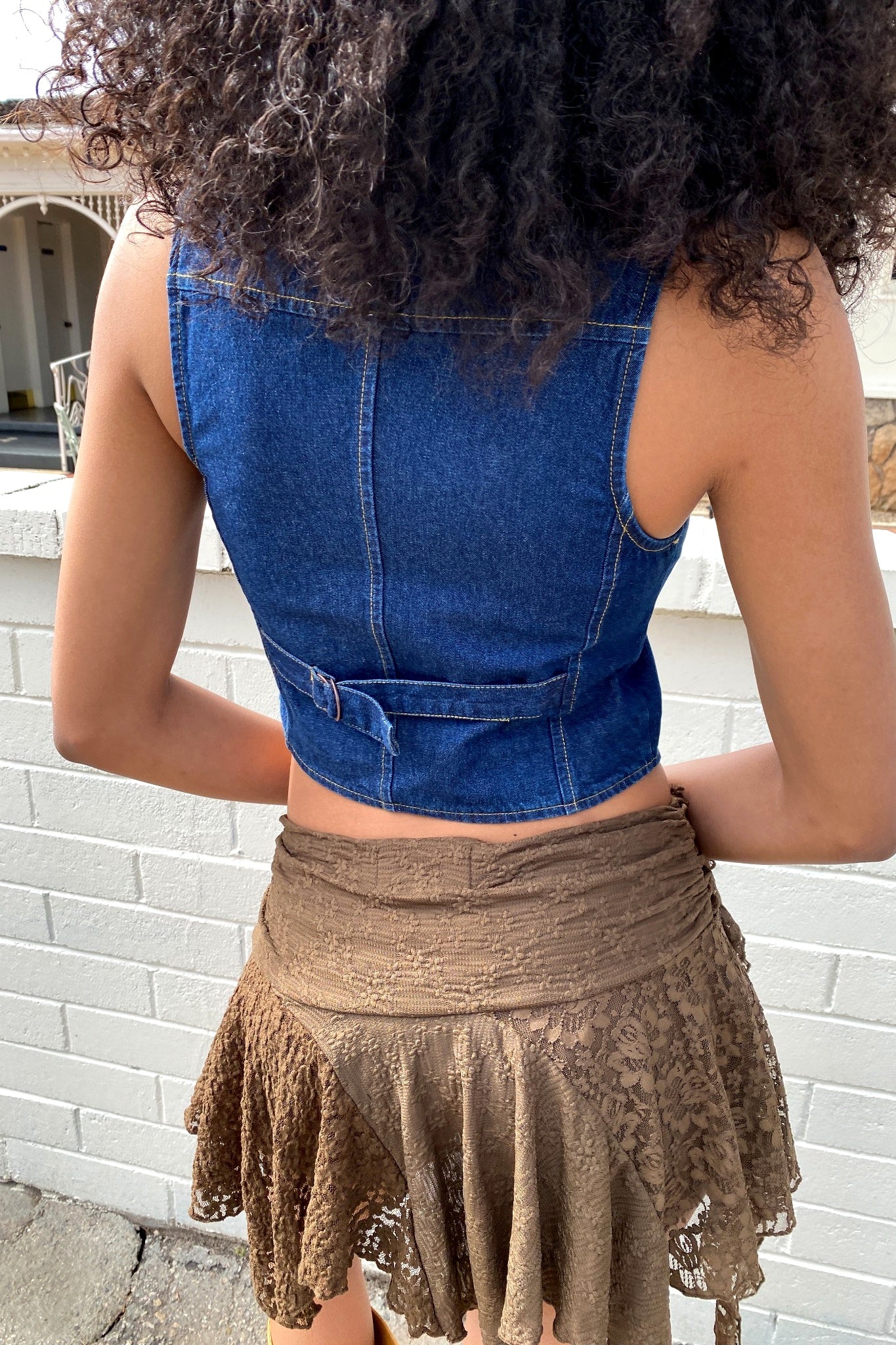 Person wearing a blue denim crop top and brown lace skirt against a white brick wall.