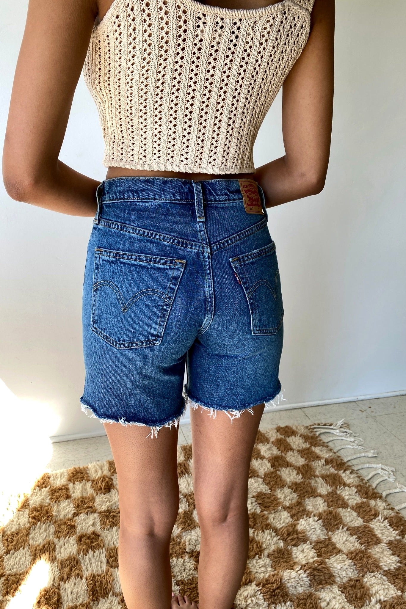 Person wearing a beige crochet top and blue denim shorts on a checkered rug.