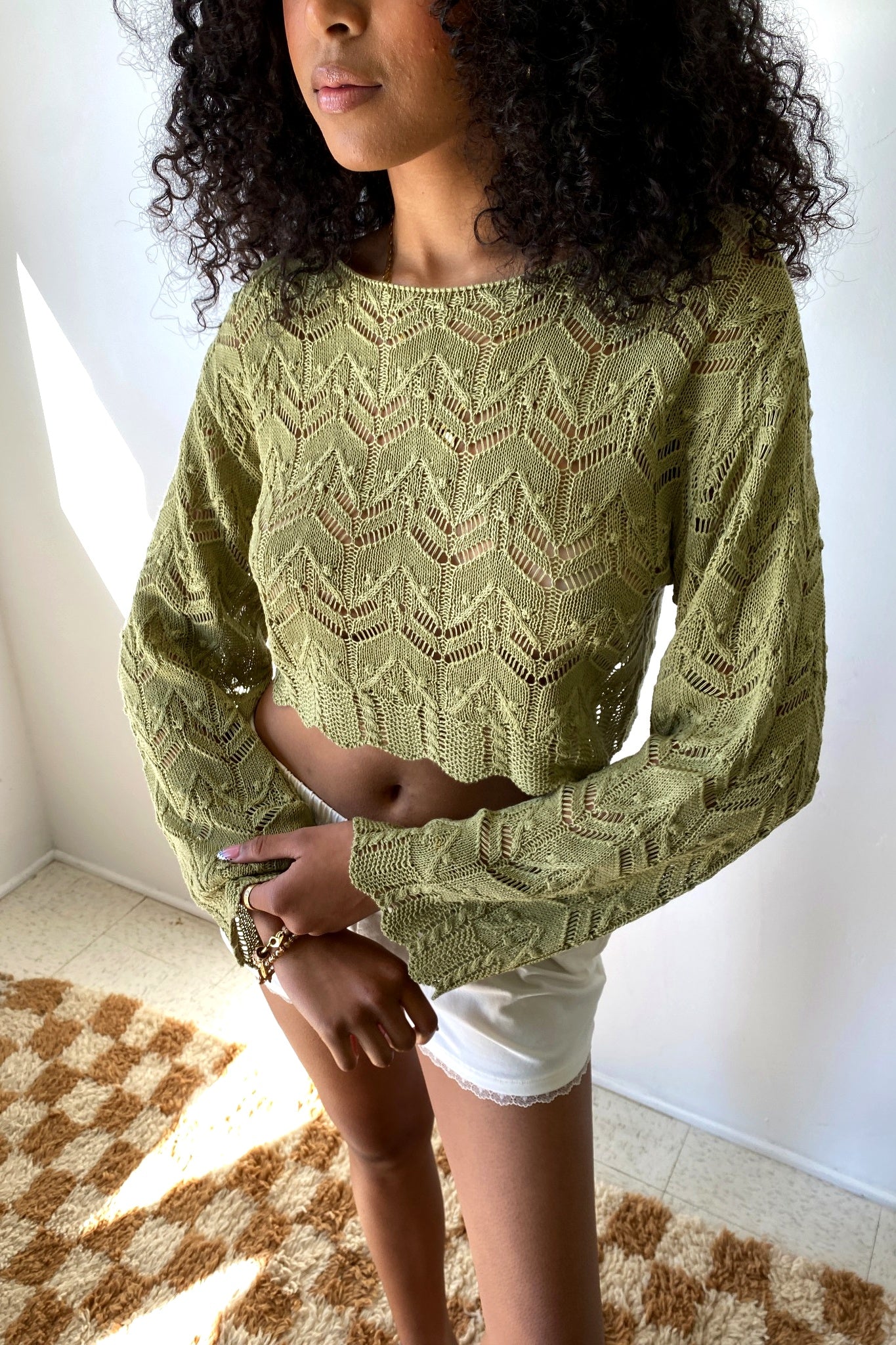 Woman wearing a green knitted sweater and white shorts in a room with a patterned rug.