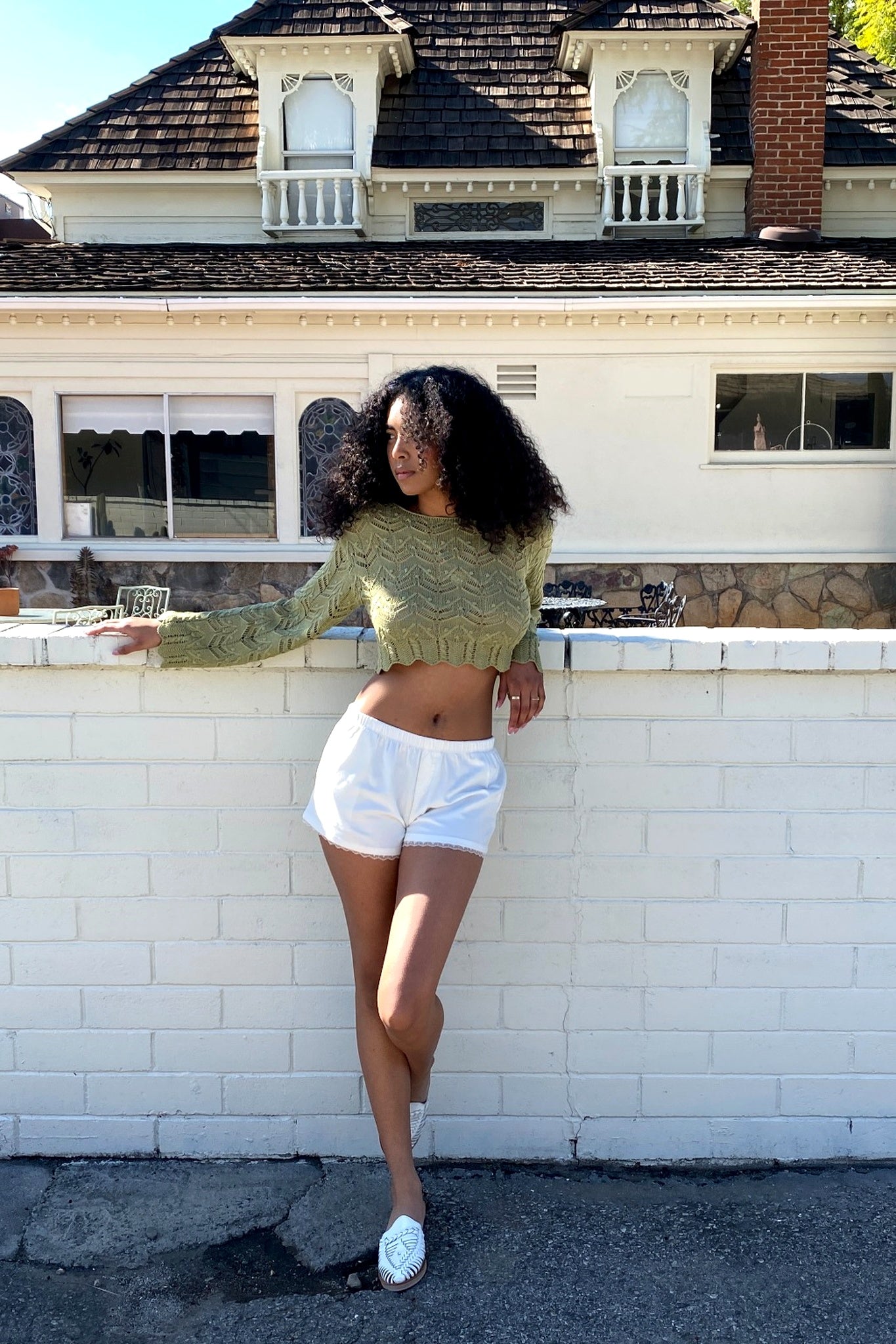 Woman in a green sweater and white shorts standing in front of a white brick wall with a house in the background.
