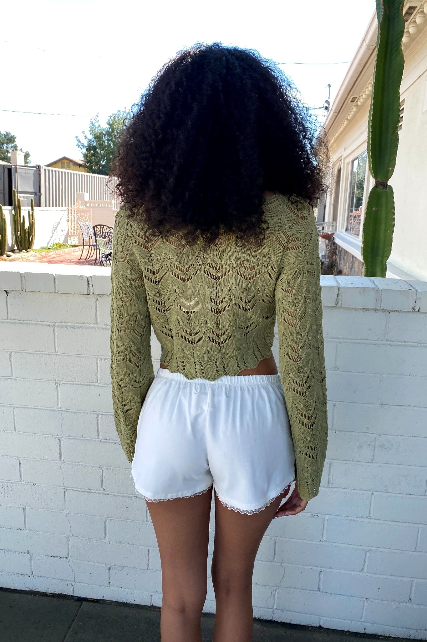 Person wearing a green knitted sweater and white shorts standing in front of a white brick wall with a cactus plant.