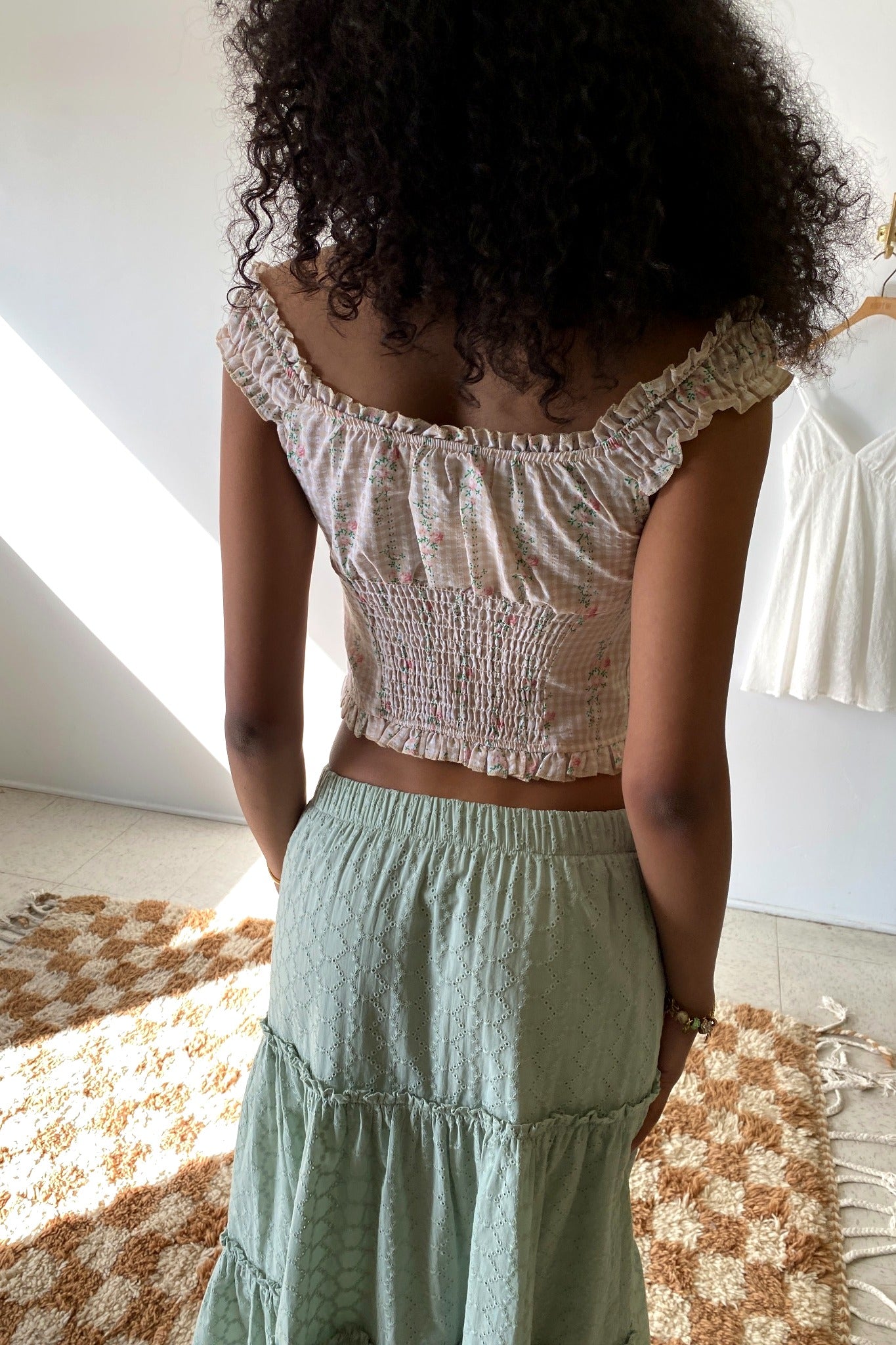 Person wearing a light-colored off-shoulder top and green skirt in a sunlit room.