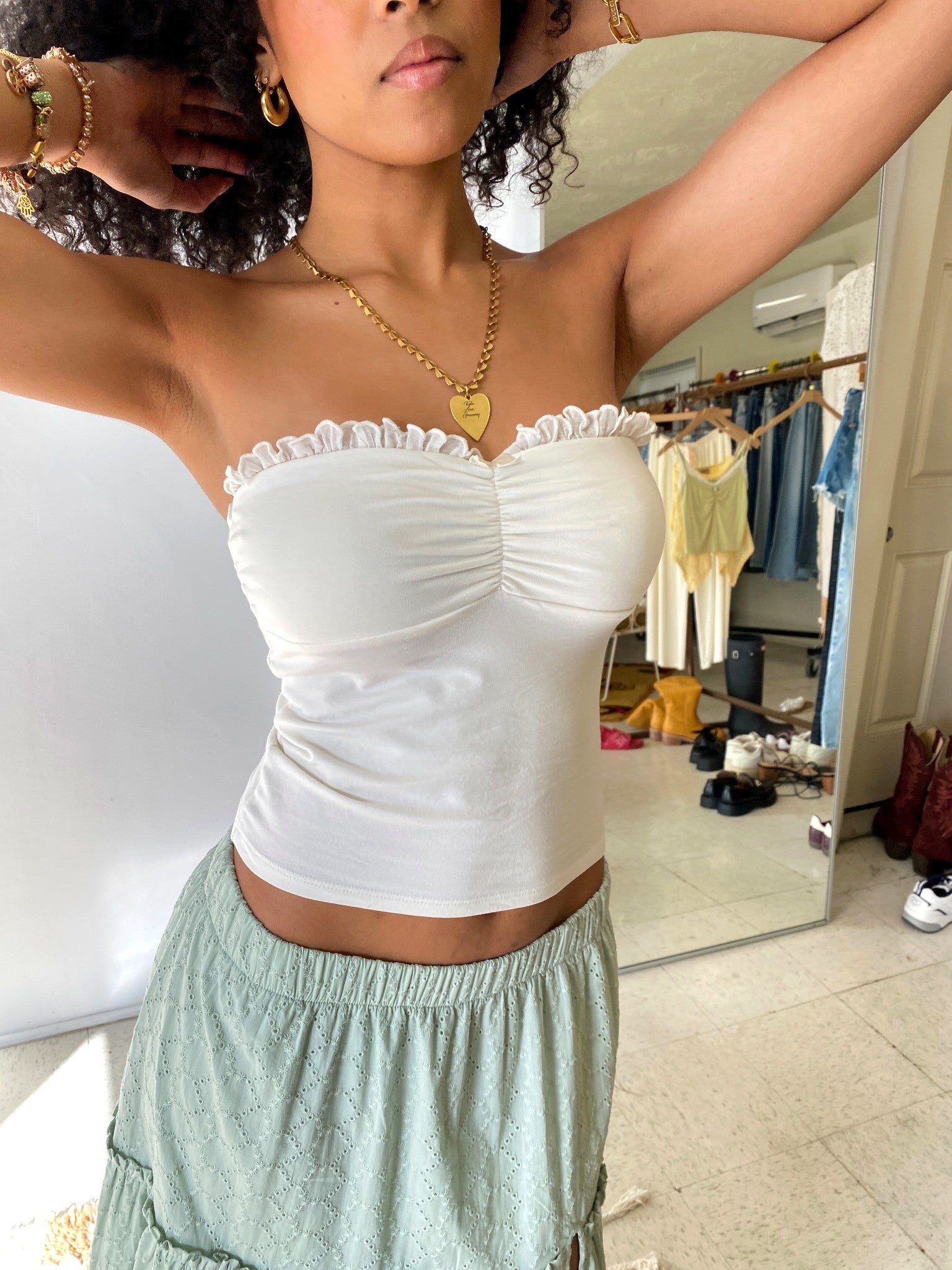 Woman wearing a white strapless top and light green pants in a room with clothing in the background.