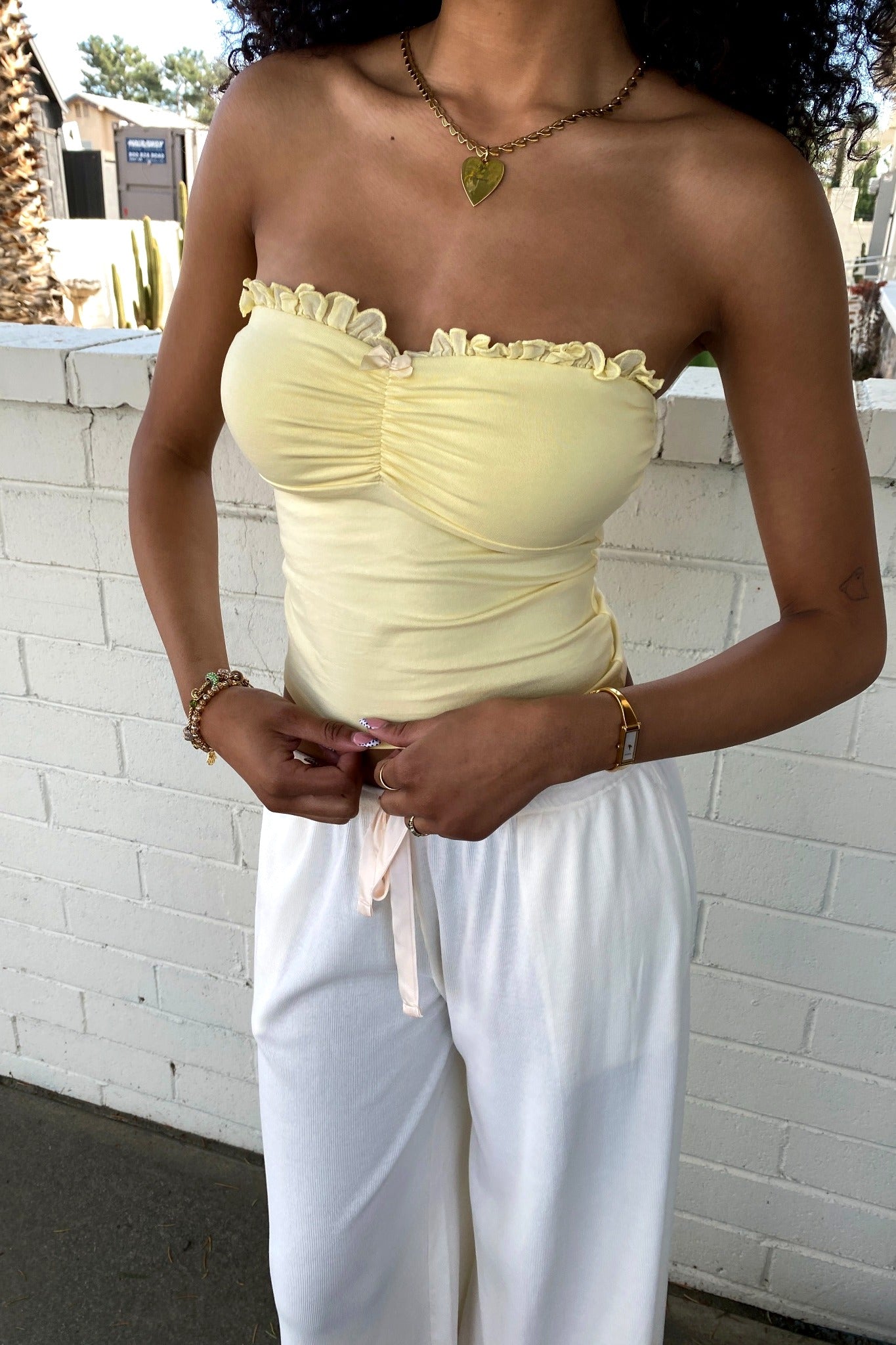 Person wearing a light yellow strapless top and white pants against a white brick wall.