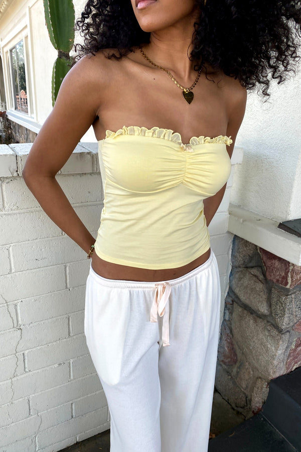 Woman wearing a yellow strapless top and white pants against a white brick wall.
