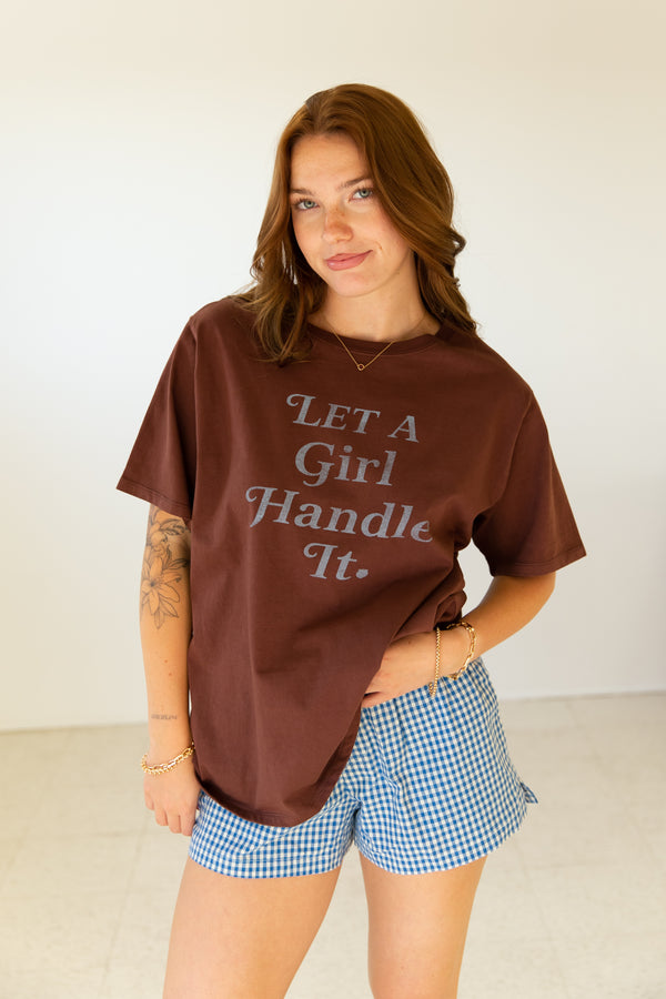 Woman wearing a brown t-shirt with 'Let a Girl Handle It' text and blue checkered shorts.