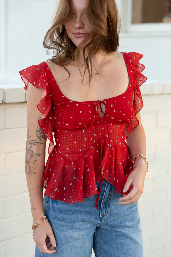 Woman wearing a red ruffled top with blue jeans against a white wall.