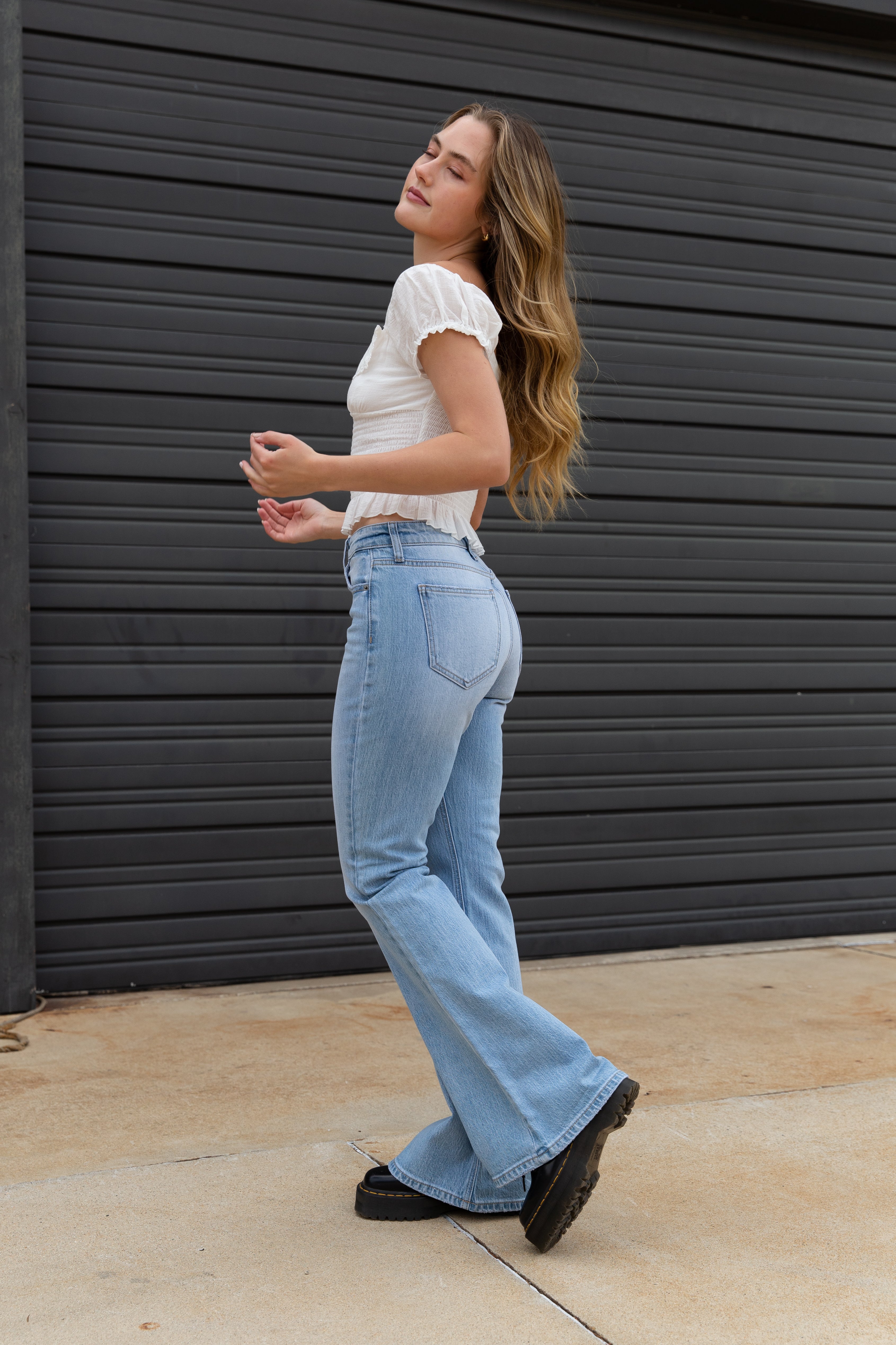 Woman wearing a white top and blue jeans standing against a dark gray wall.