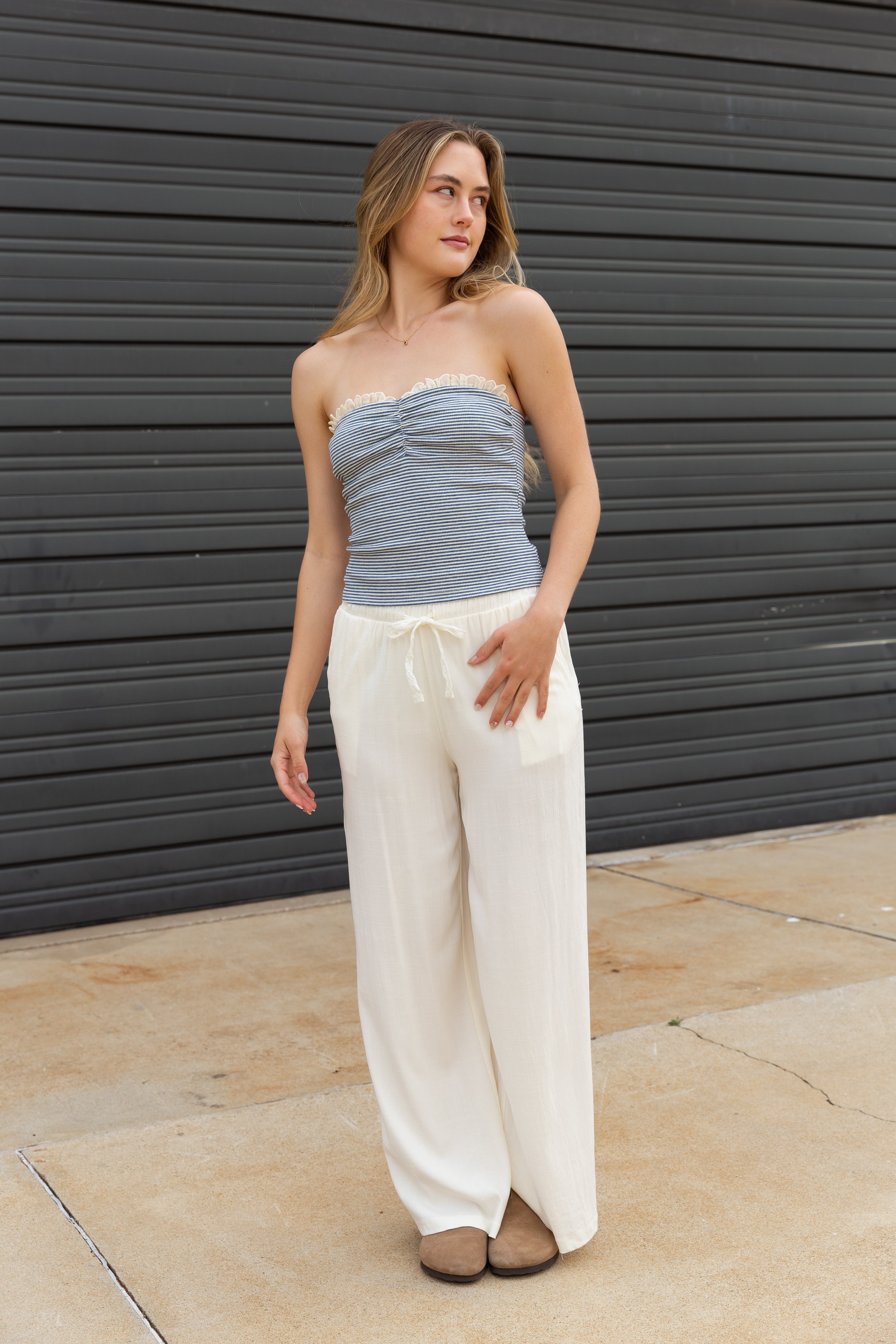Woman wearing a strapless top and wide-leg pants standing against a dark wall.