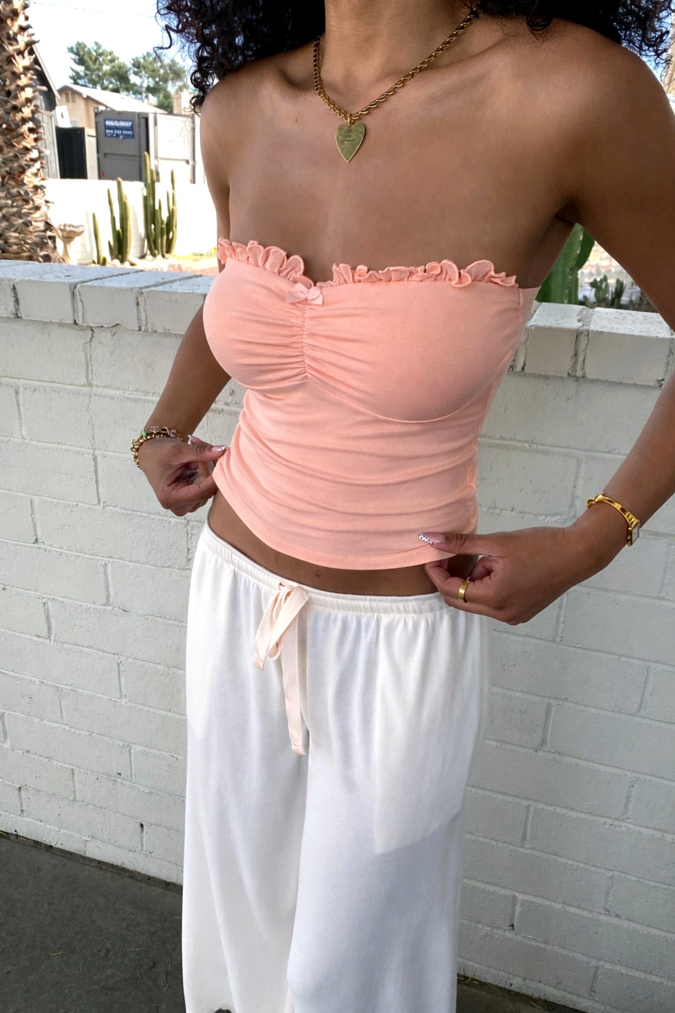 Person wearing a pink strapless top and white pants against a white brick wall.