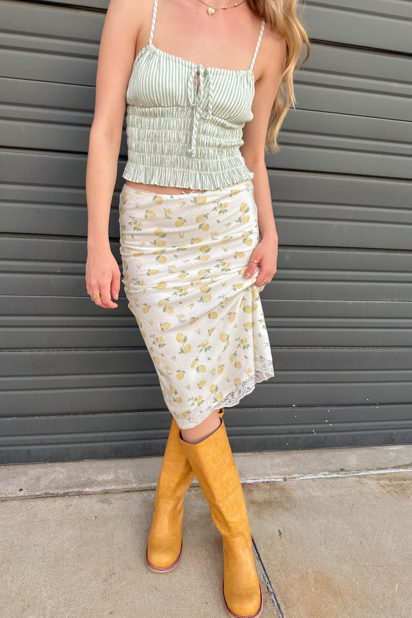 Person wearing a light green top and white floral skirt with yellow boots against a gray wall.