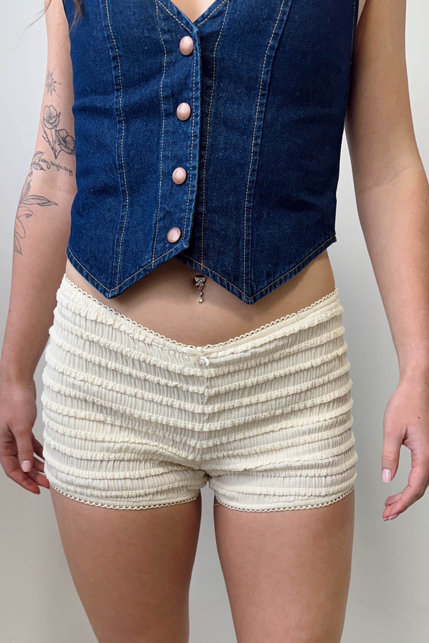 Person wearing a denim vest over a white top and beige shorts on a plain background