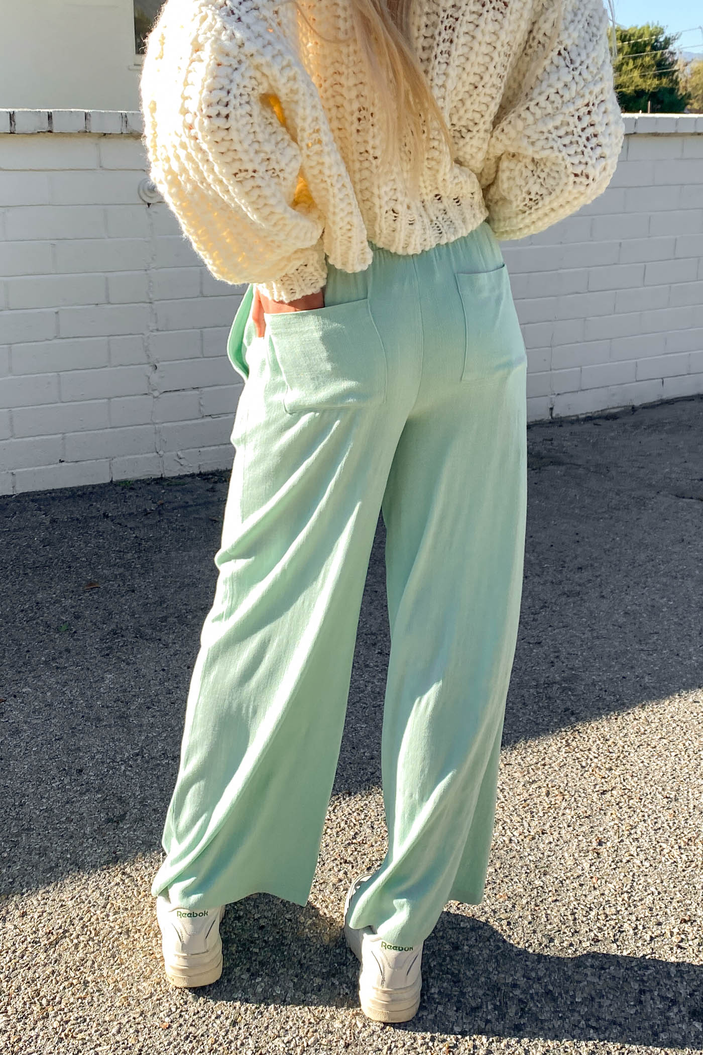 Person wearing a light-colored sweater and light green pants standing on a street.