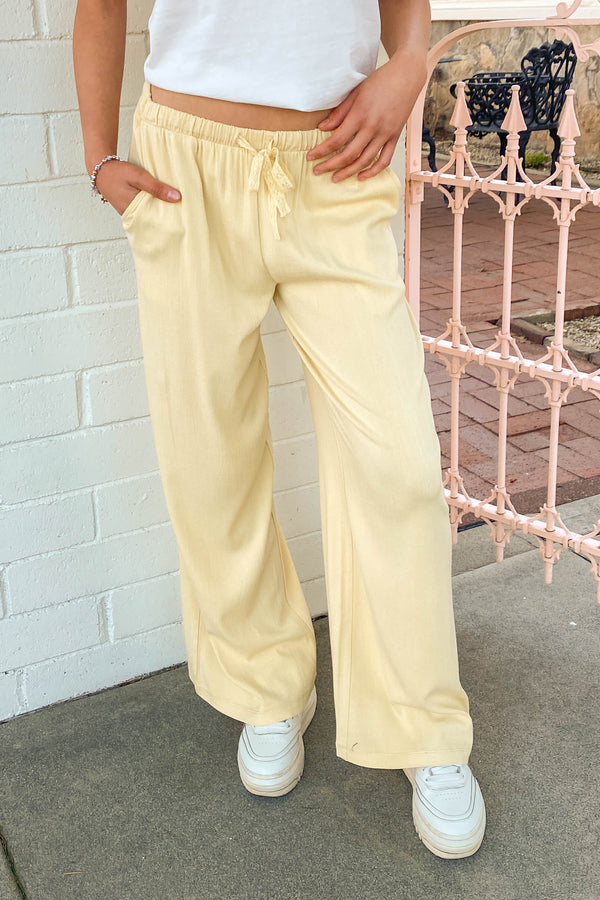 Person wearing light yellow pants and white sneakers standing against a white brick wall.
