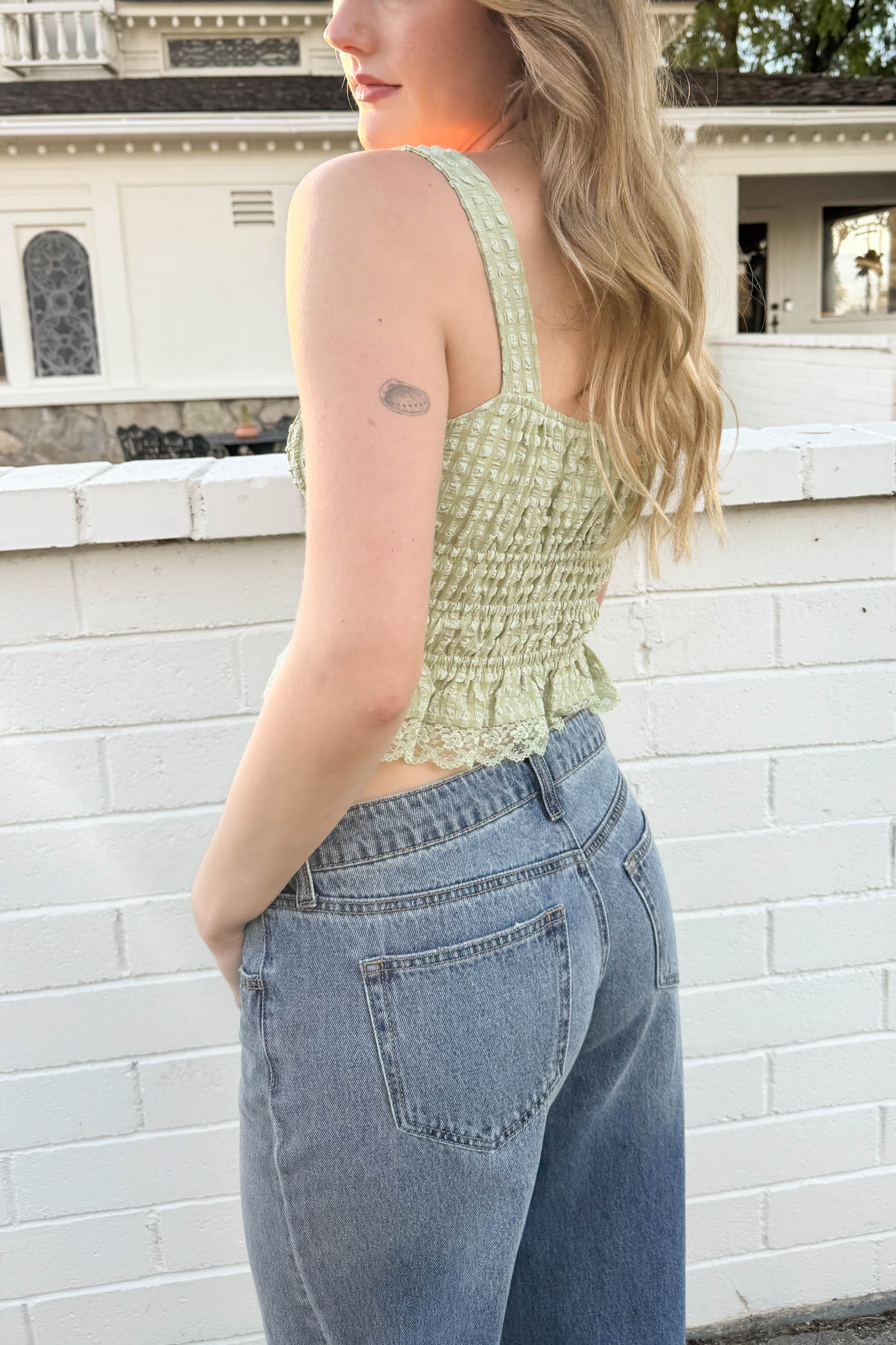 Woman wearing a green tank top and blue jeans against a white brick wall.