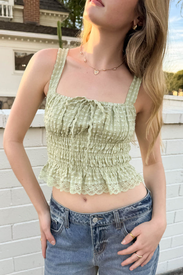 Woman wearing a green lace top and blue jeans against a white brick wall.