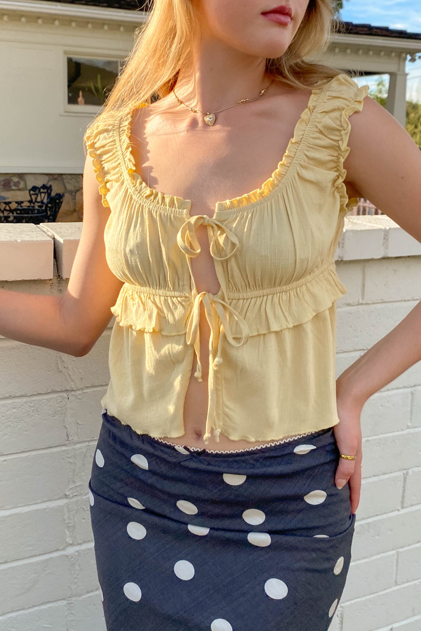 Woman wearing a yellow top with ruffles and a navy skirt with white polka dots, standing against a white brick wall.