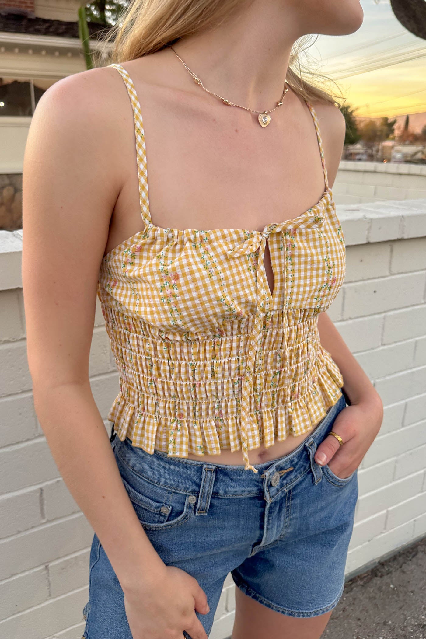 Woman wearing a yellow checkered top and denim shorts against a white brick wall.