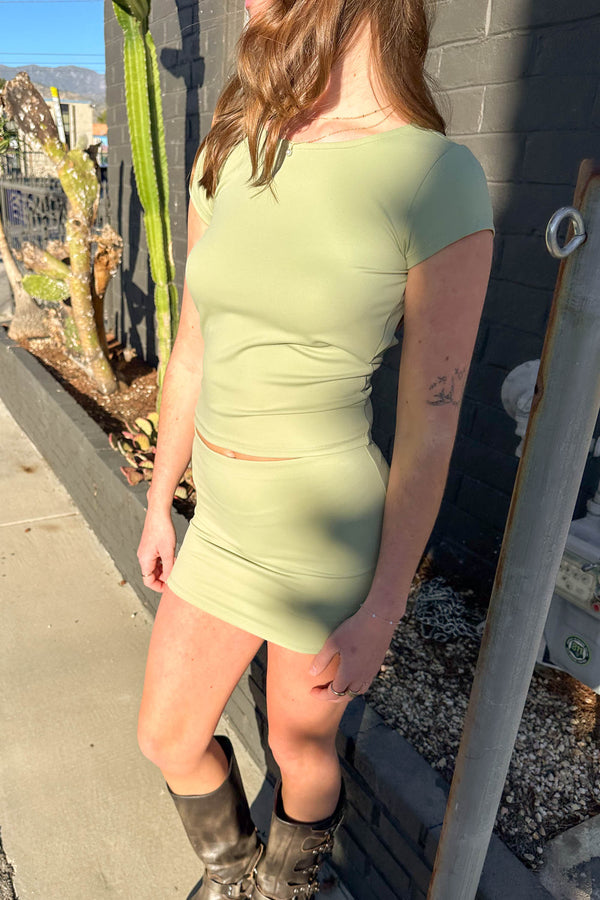 Person wearing a light green dress standing against a wall with plants in the background