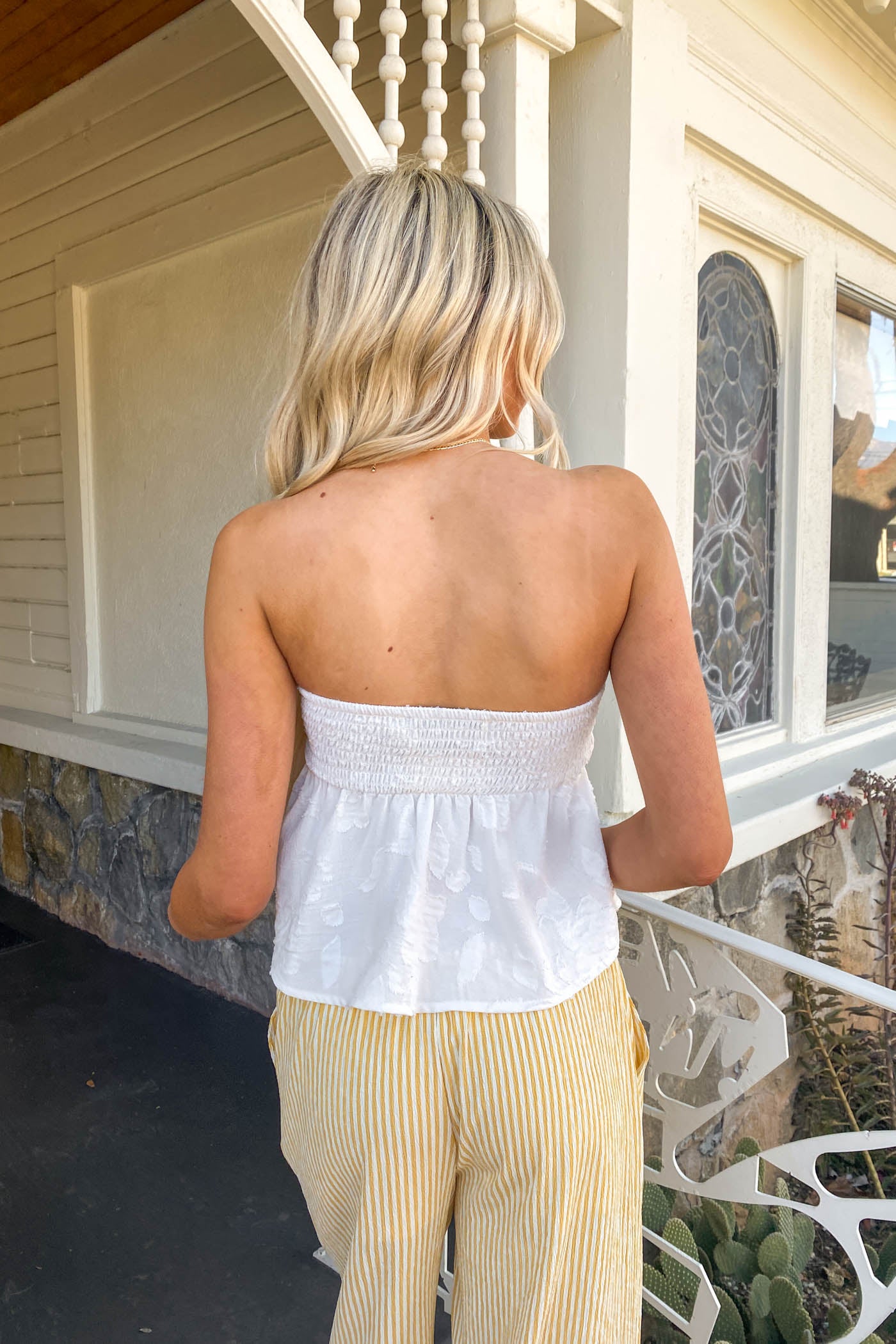 Woman wearing a white strapless top and beige pants standing on a porch.