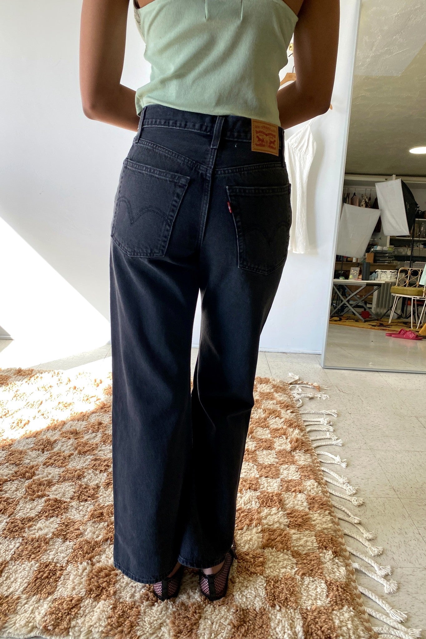 Person wearing dark blue jeans and a light green top in a room with a mirror and furniture.