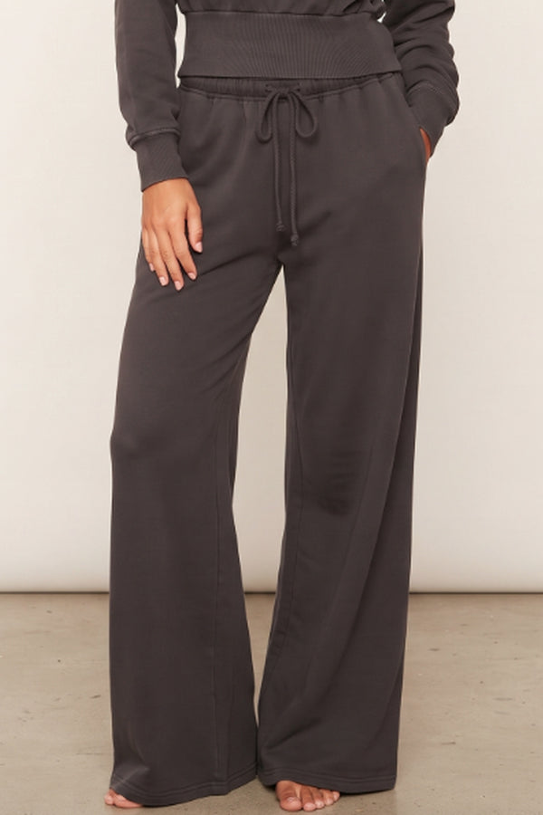 A person wearing dark charcoal wide-leg sweatpants with a drawstring waist and side pockets, paired with a matching dark sweatshirt, shown from the waist down against a plain light wall.